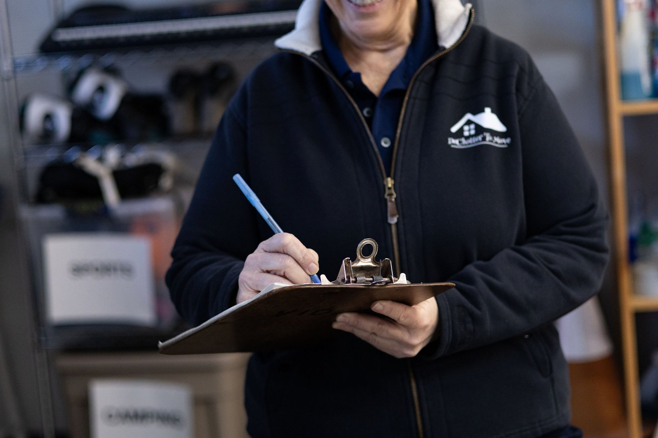 teri webster professional organizer  in a dark jacket writing on a clipboard with a pen inside a workshop or storage room.