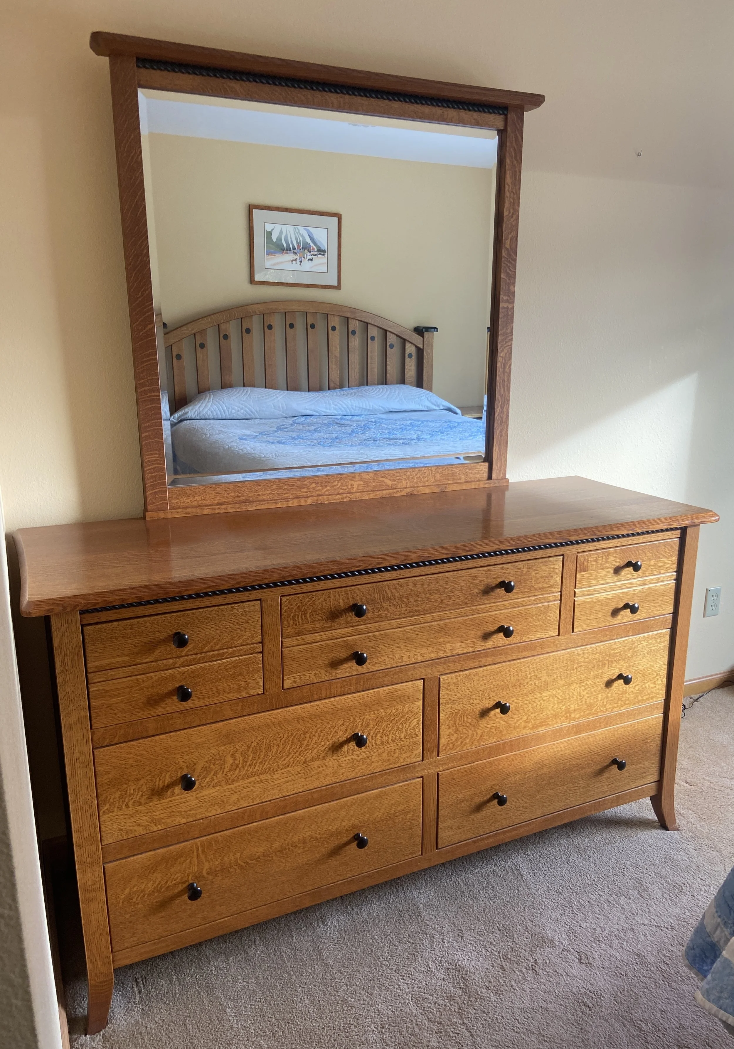 Wooden dresser with a mirror reflecting a bed and a picture on the wall.