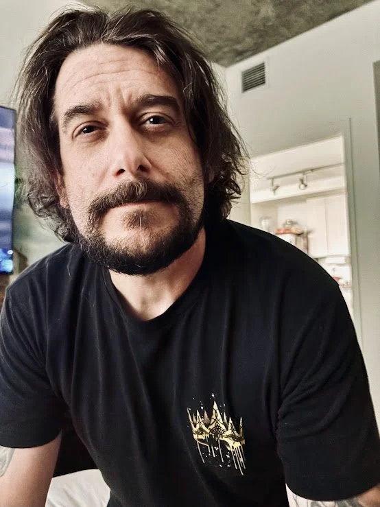 A man with long brown hair and a beard, wearing a black T-shirt, looking at the camera in a room with a TV and kitchen in the background.