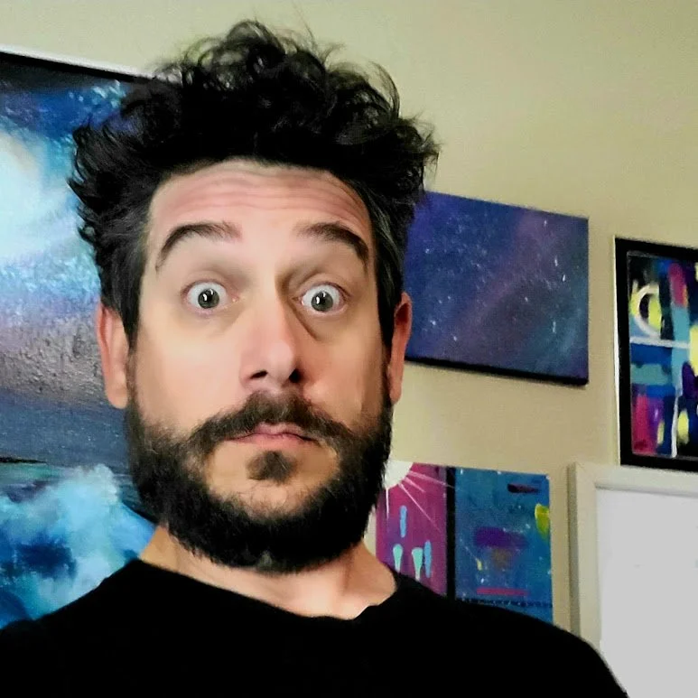 A man with dark, curly hair and a beard, making a surprised facial expression in front of colorful abstract artwork.
