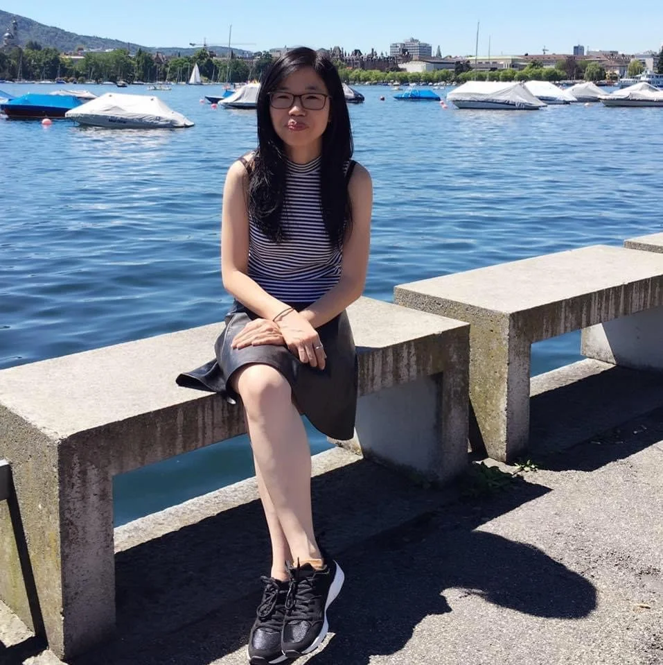 A woman with glasses and long black hair, sitting on a concrete bench by a body of water on a sunny day, with boats and a city skyline in the background.