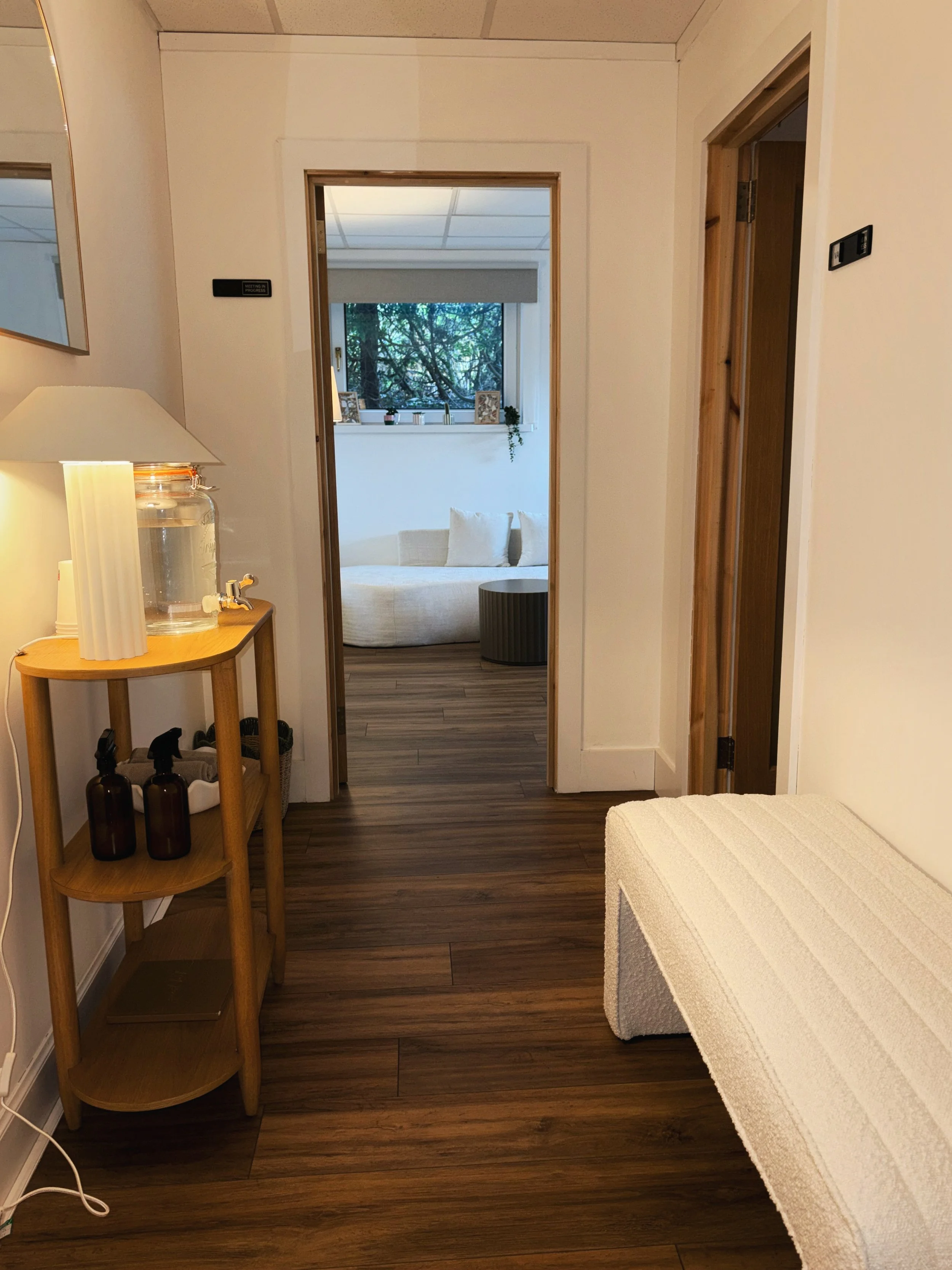 A cozy room hallway with a wooden floor, a small side table with bottles and a water dispenser, a white textured bench, and a view into a living room with a curved sofa and a window with trees outside.