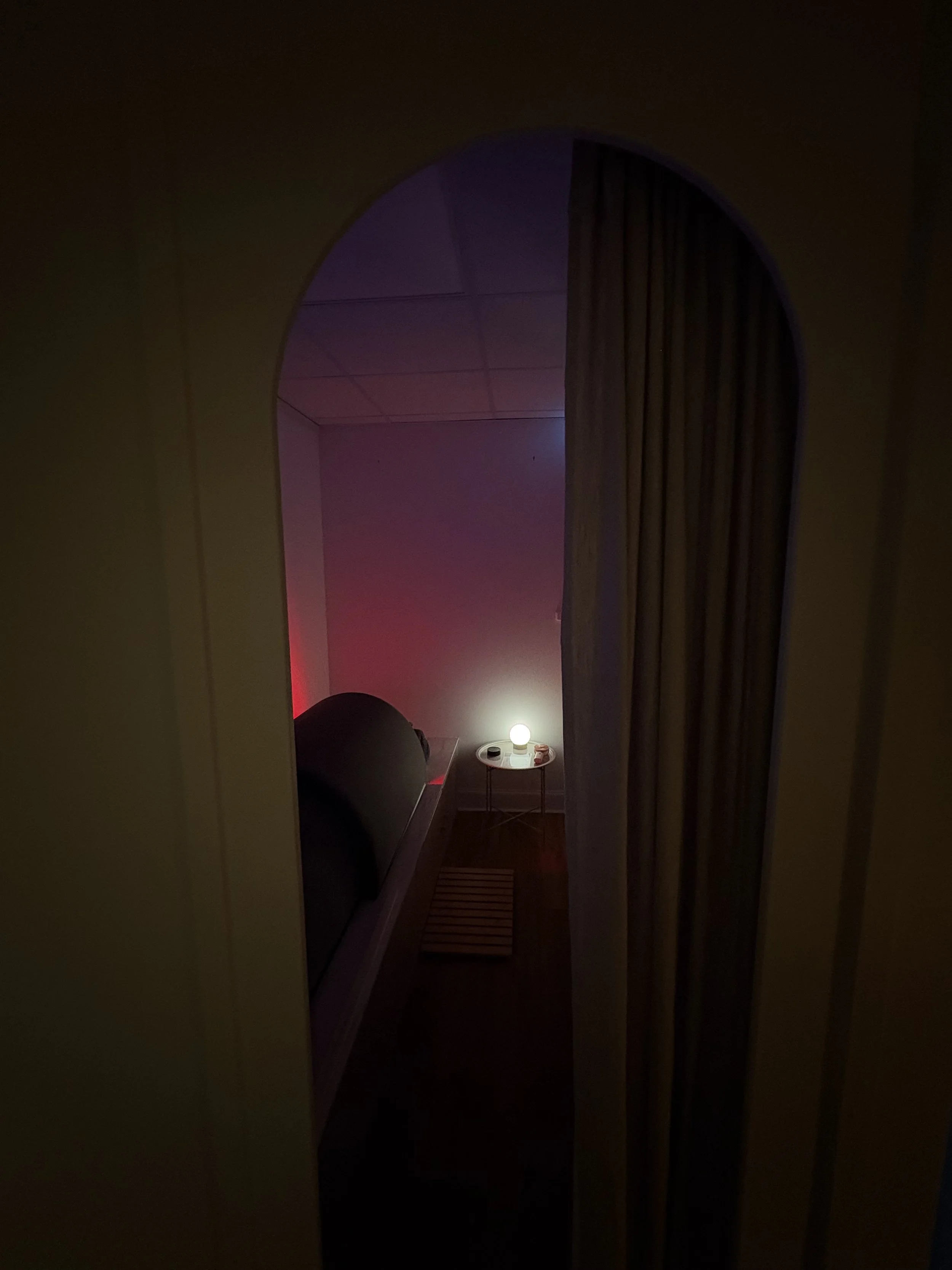 Dark view of a bedroom seen through an archway, with a small bedside table holding a glowing lamp, a bed, and some items on the table.
