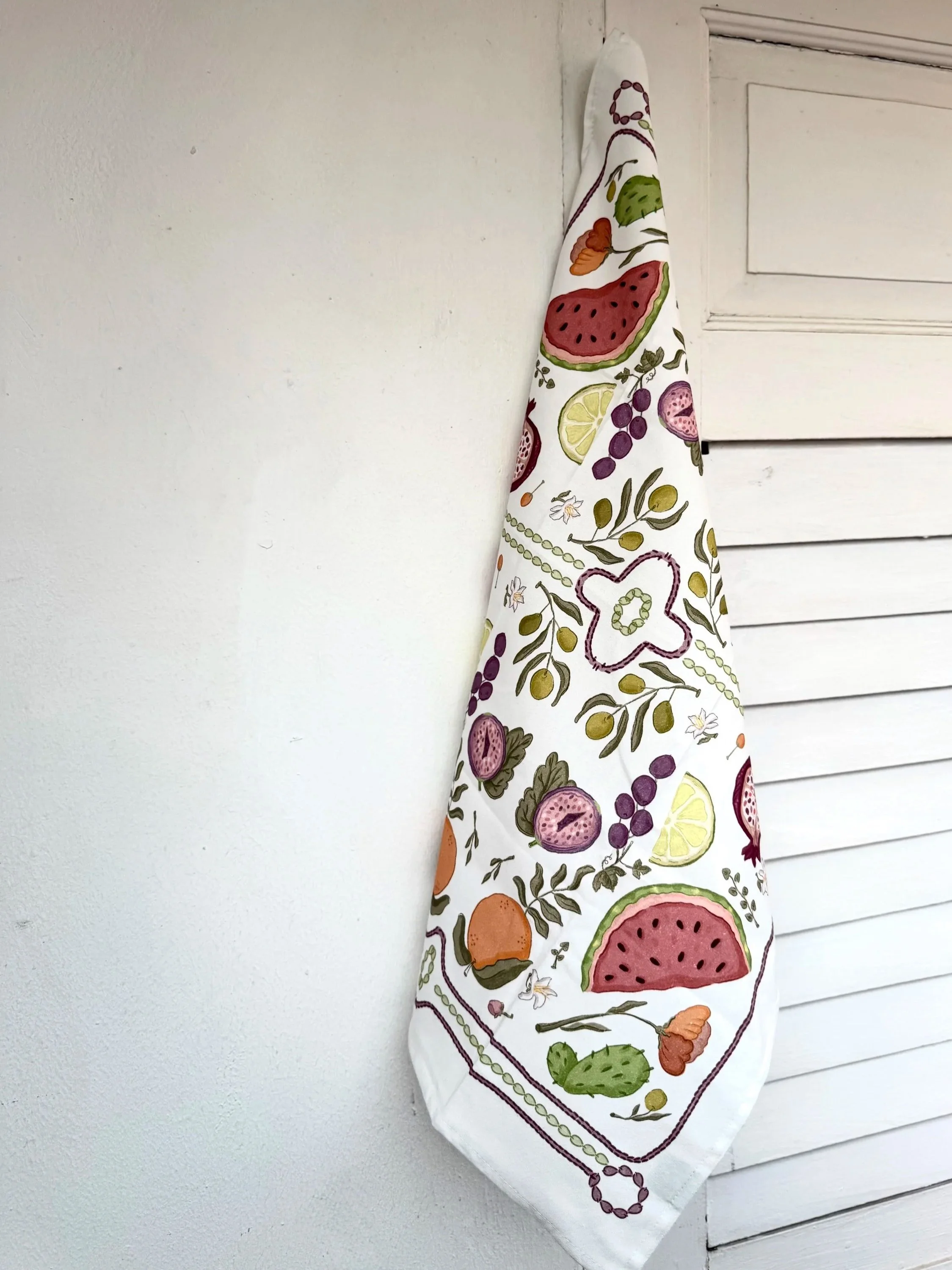 A white kitchen towel with colorful fruit and floral patterns hanging on a wall next to a white louvered door.