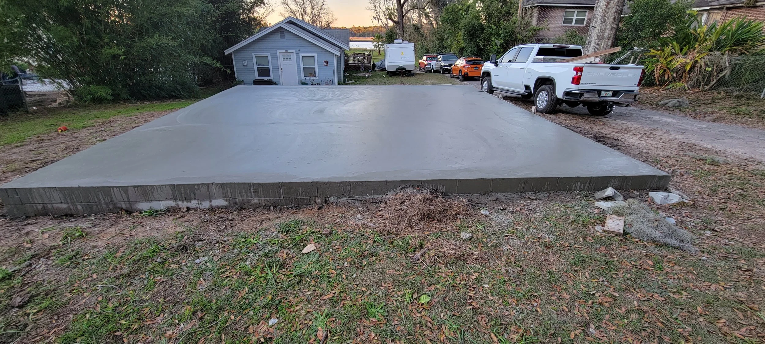 A freshly poured concrete slab with a brick border, situated in a backyard with a small blue house, several parked cars, and trees in the background, during early evening.