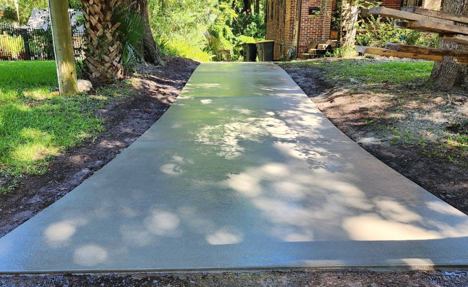 Newly poured concrete sidewalk in a residential yard, with shadows from trees cast across the surface, leading towards a brick house in the background.