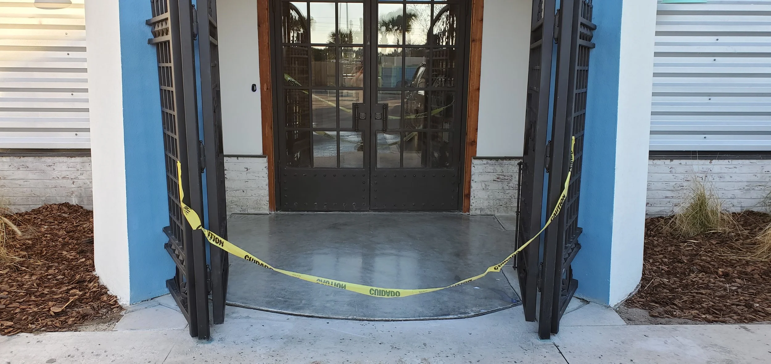 Entrance with double glass doors surrounded by black metal gates, caution tape across the entrance, white wall with blue accents, dirt mulch on each side of the walkway.