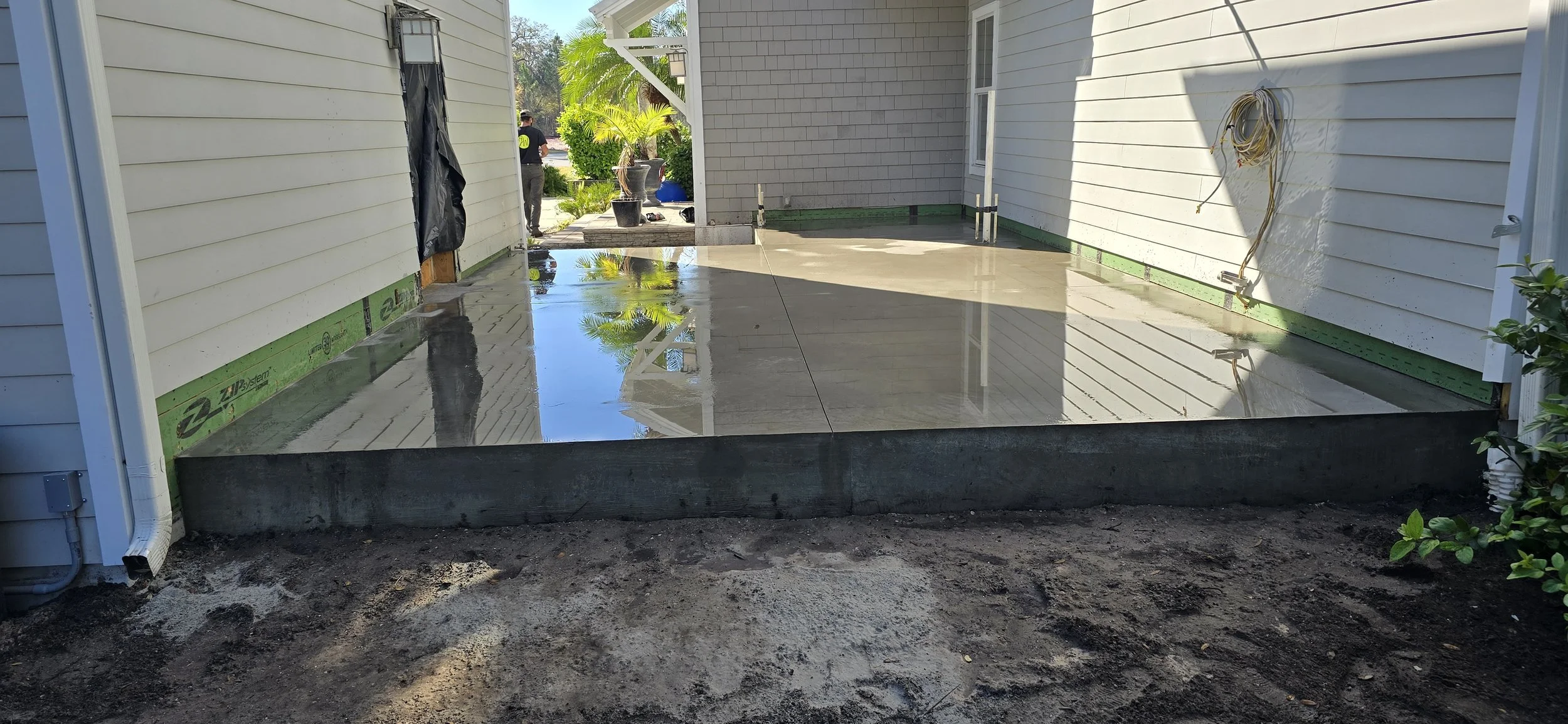 Freshly poured concrete patio between two houses with white siding, with some construction supplies and a person in the background, and reflecting the house and sky in the wet surface.