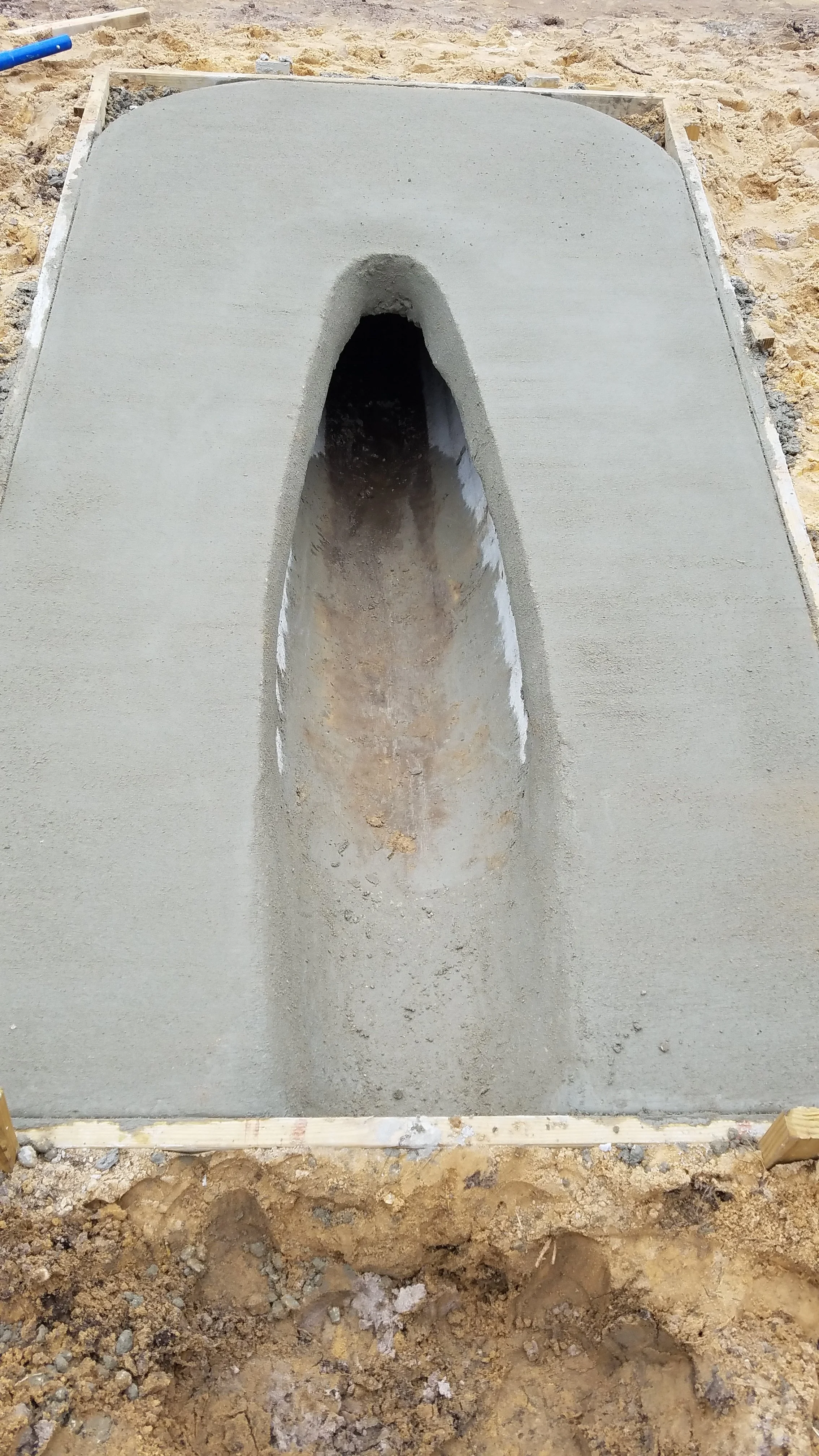 A concrete foundation with a vertical, elongated oval-shaped cutout in the center, surrounded by sandy soil and framing wood.