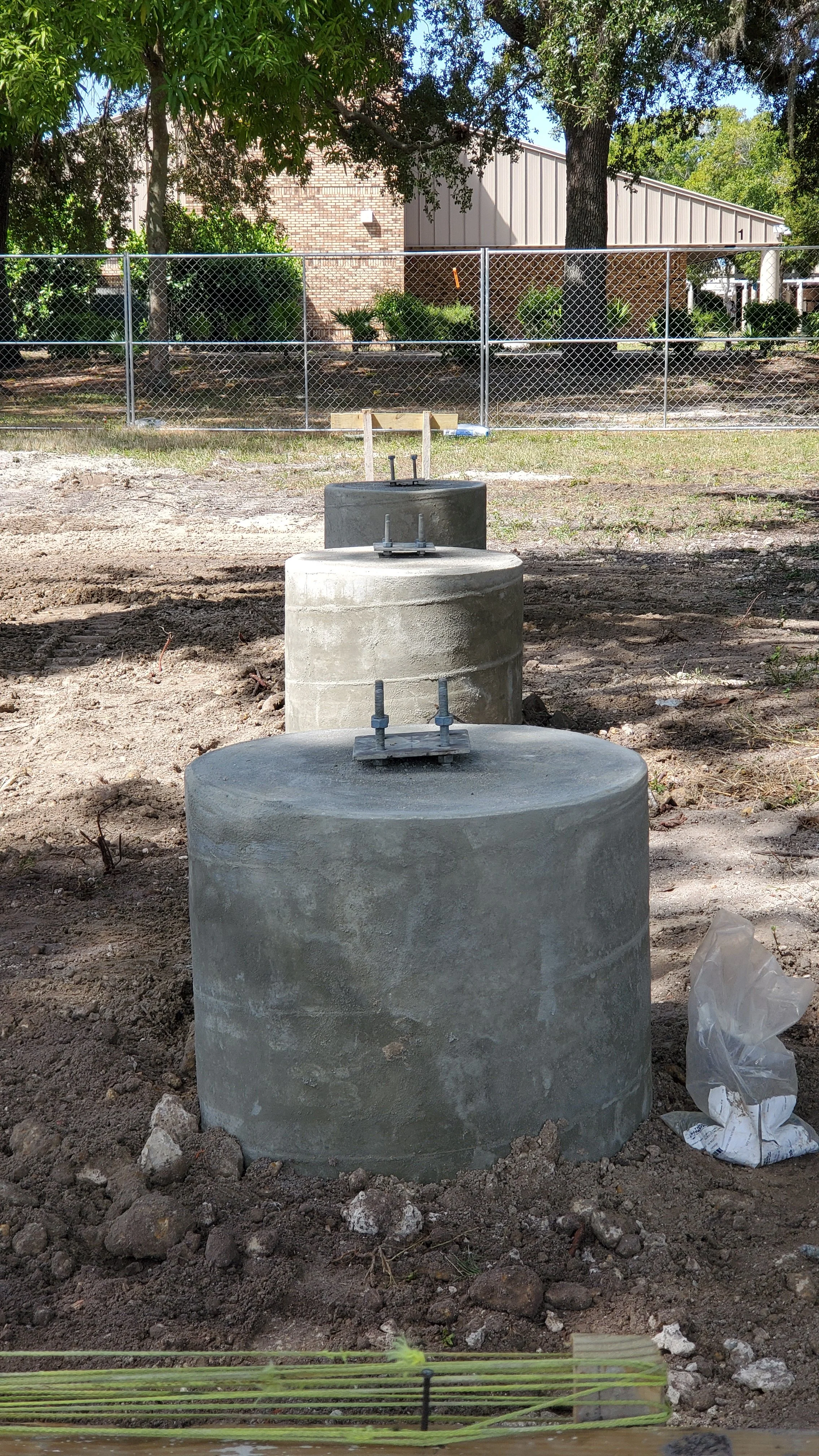 Three concrete foundation pillars with metal bolts on top, set in soil, on a construction site near a fence and trees.