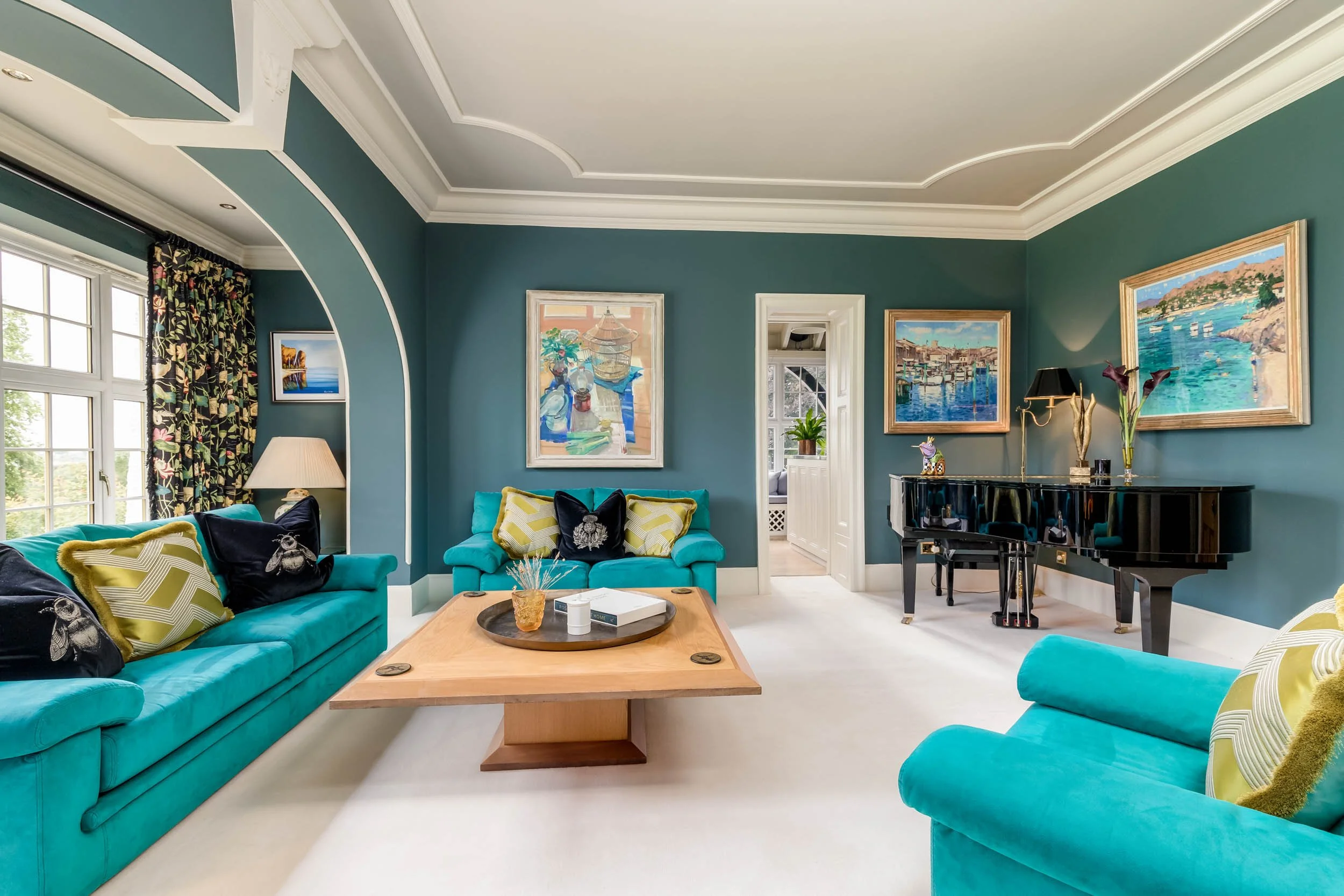 Living room with teal sofas, a wooden coffee table, a black grand piano, and colorful framed landscape paintings on teal walls.