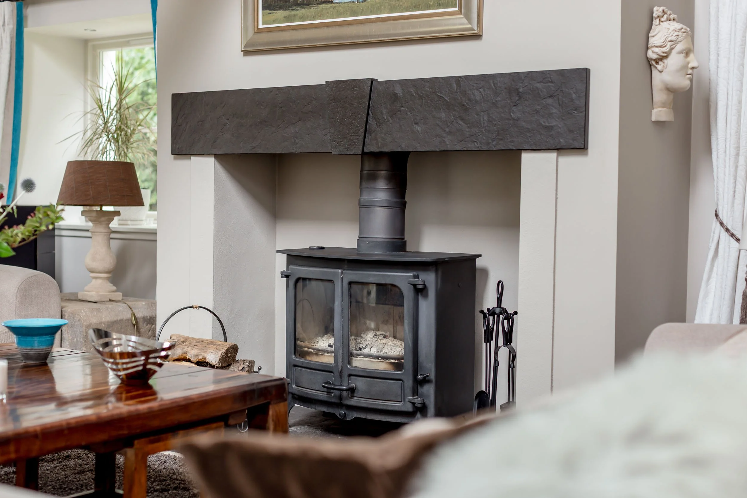 Living room with a wood-burning stove set in an alcove, with a black metal chimney and firewood nearby. Decor includes a bronze table lamp, a framed painting, and a wall sculpture of a classical woman's bust.
