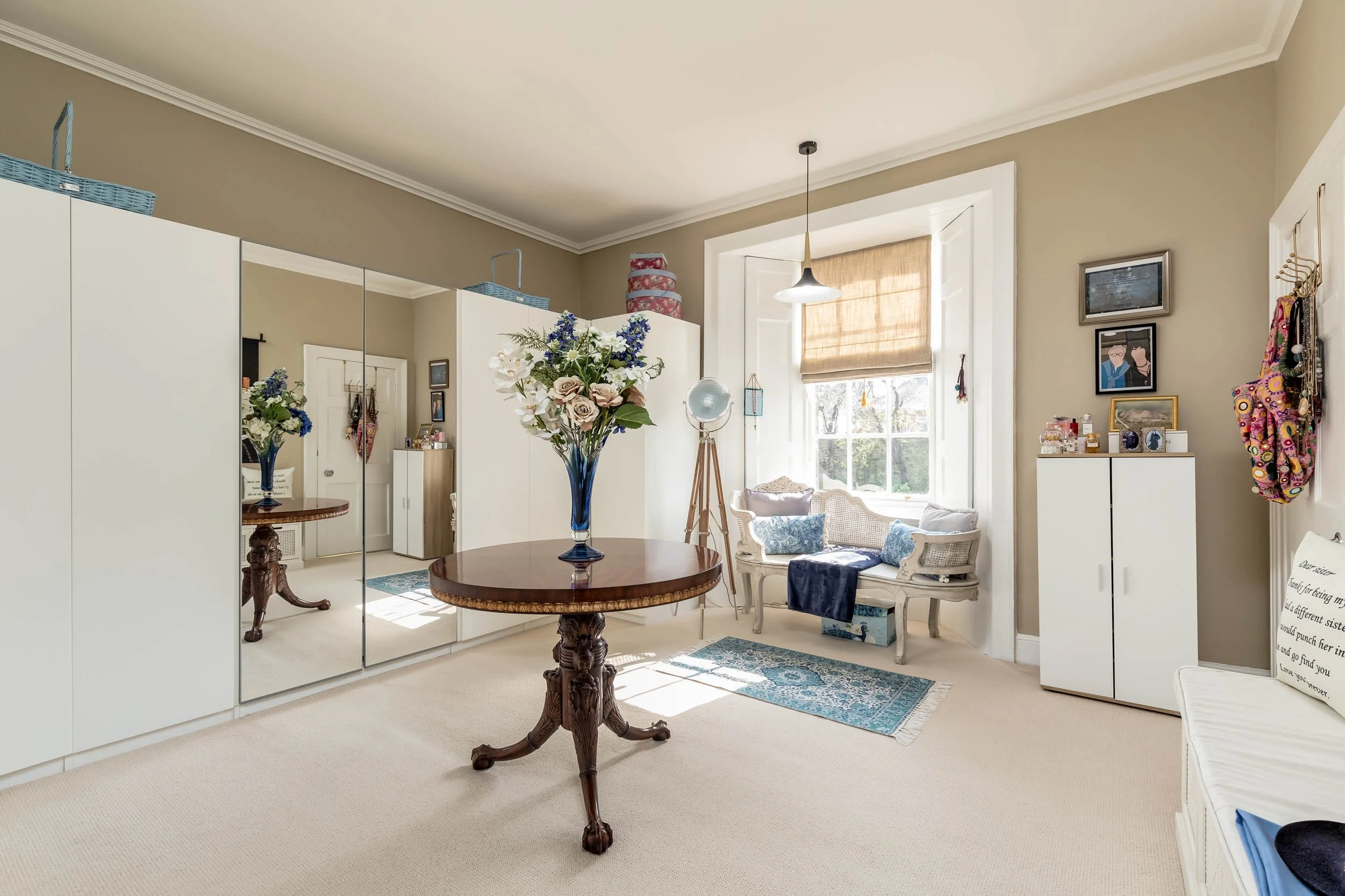 Living room with beige walls, carpeted floor, white storage cabinets, a round wooden table with a vase of flowers, a window with a beige roller shade, cushioned seating by the window, framed art on the wall, and decorative items on cabinets.