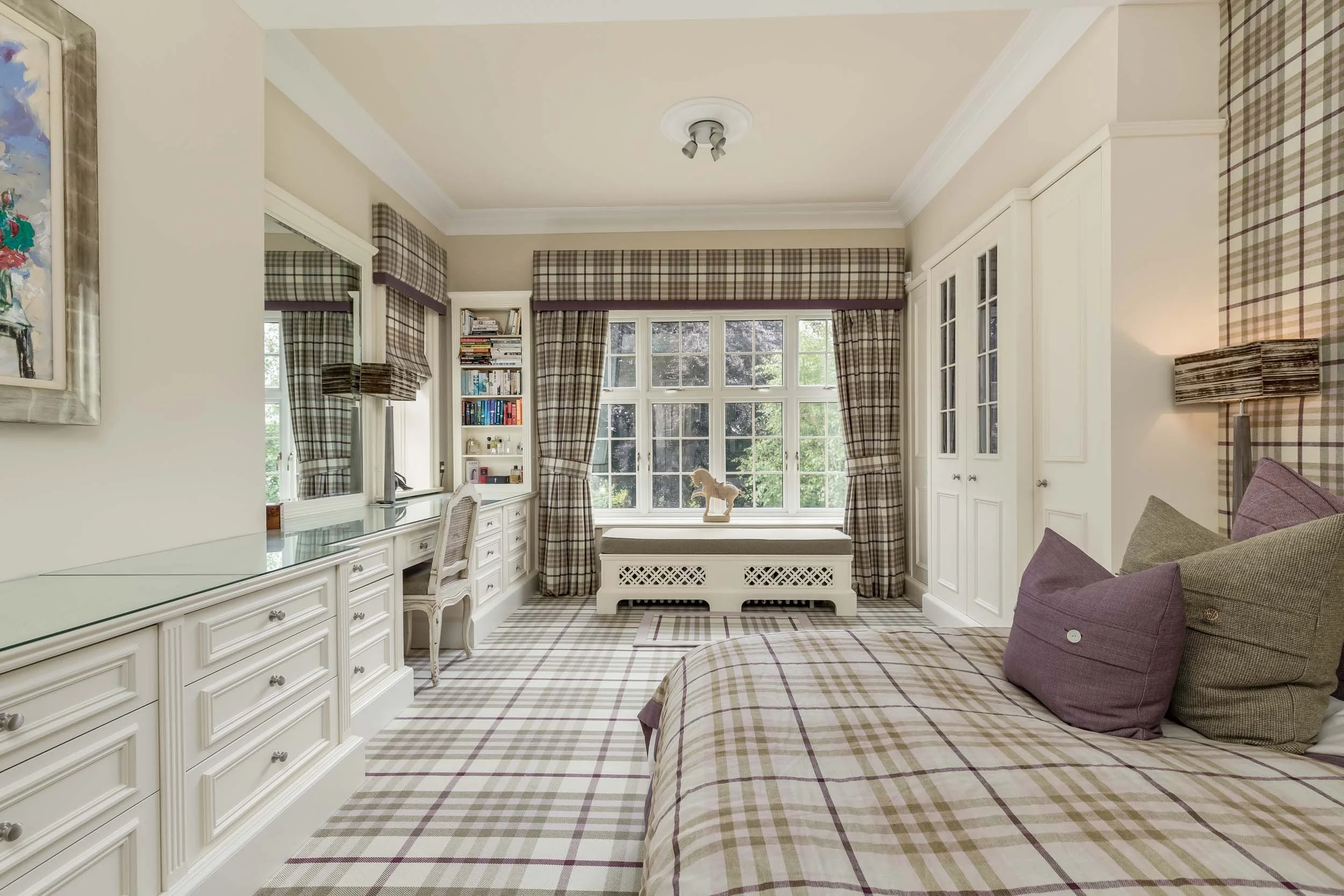 A bedroom with plaid-patterned curtains, matching carpet, and bedding, white built-in furniture, a window seat with a horse figurine, a desk with books, and purple and green pillows on the bed.
