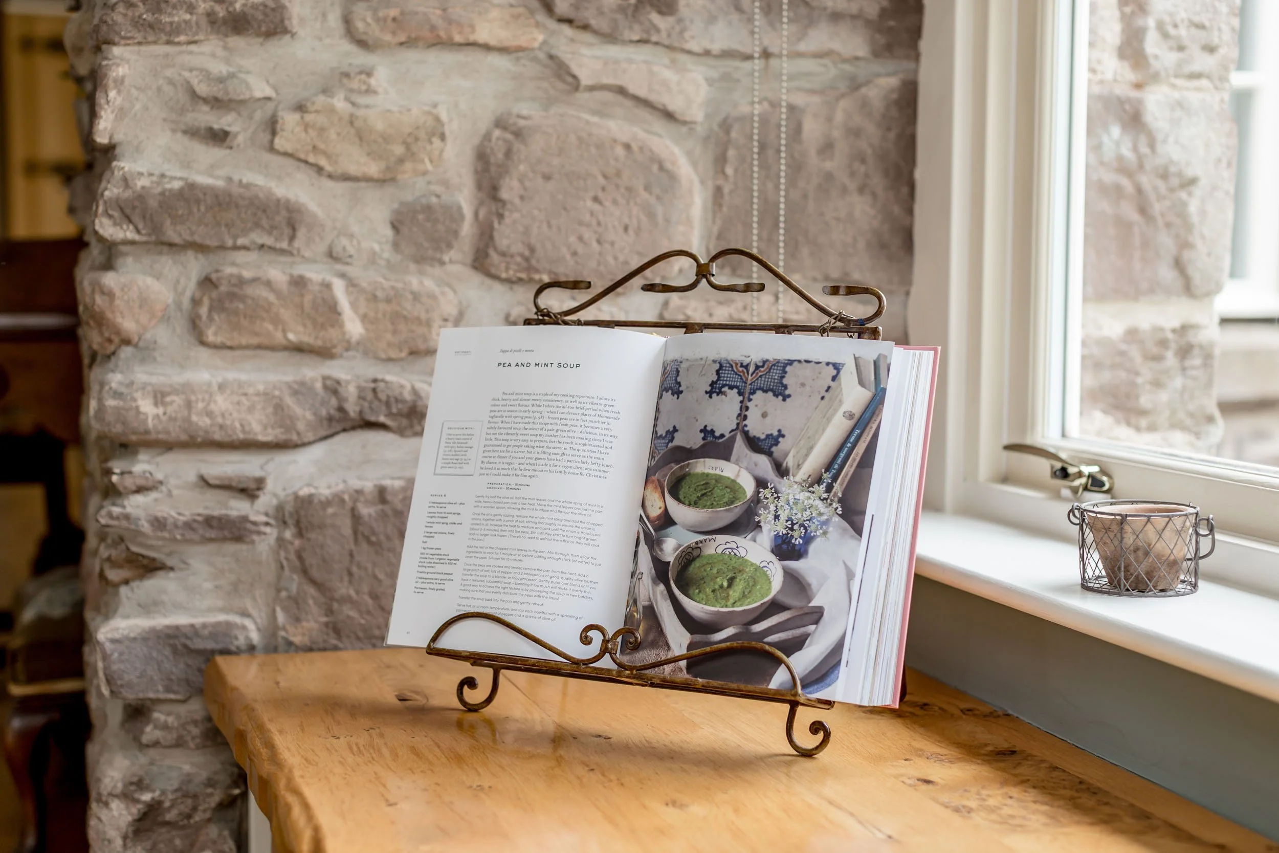 Open cookbook on a metal bookstand on a wooden table near a window with a stone wall in the background, showing a recipe for pea and mint soup and a photograph of two bowls of green pea soup.