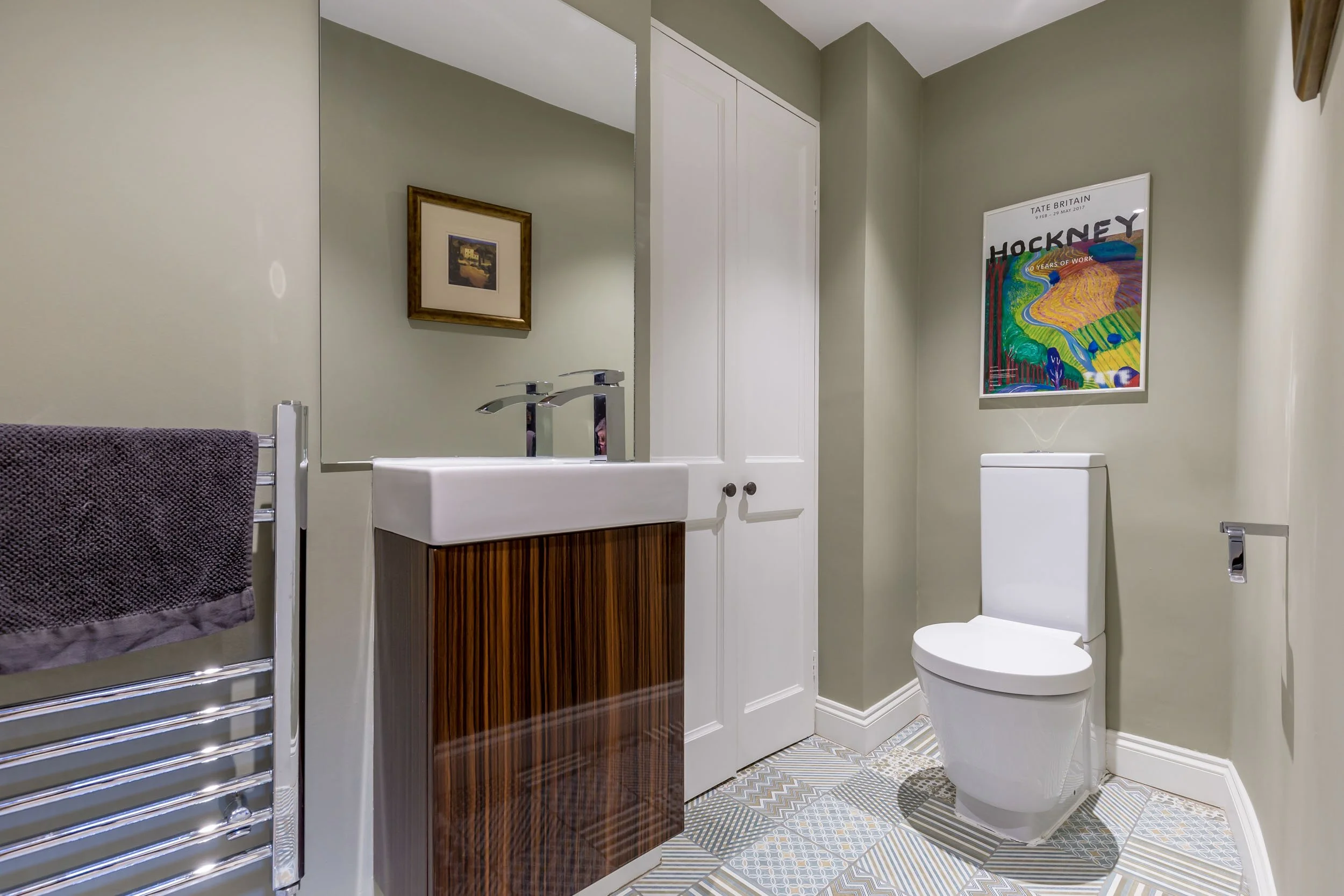Modern bathroom with a white toilet, wooden vanity with a white sink, a large mirror, framed art on the wall, and patterned tile flooring.