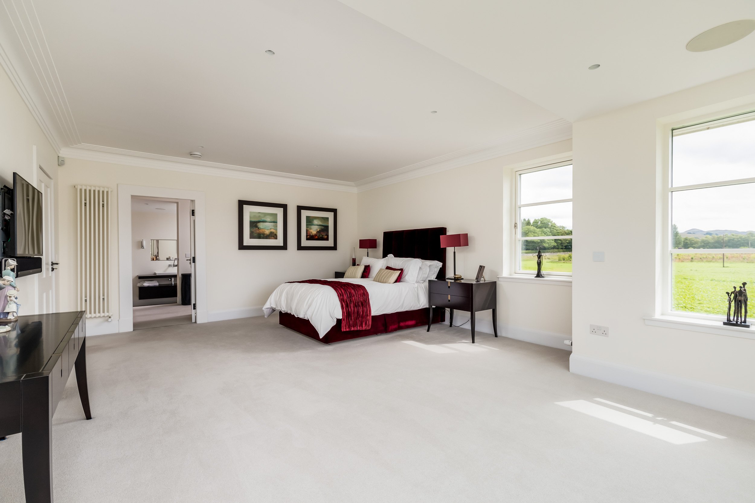 A spacious, bright bedroom with a large bed, two black nightstands with red lamps, and two framed landscape paintings on the wall. There are large windows showing a green field outside.