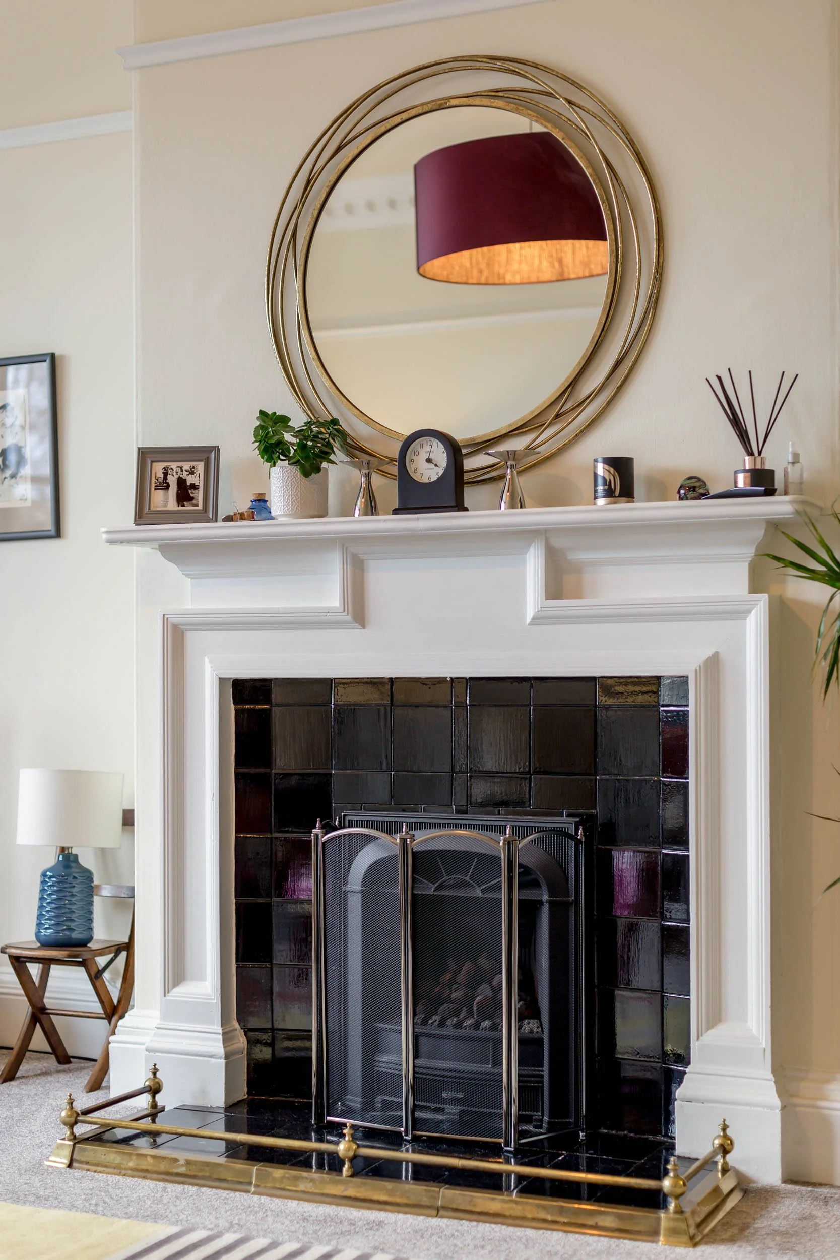 Living room fireplace with white mantel, black tiles, and a brass fireguard, decorated with a large round gold mirror, potted plant, clock, and various decorative objects on the mantel. To the left, a small side table with a blue vase and white lamp,