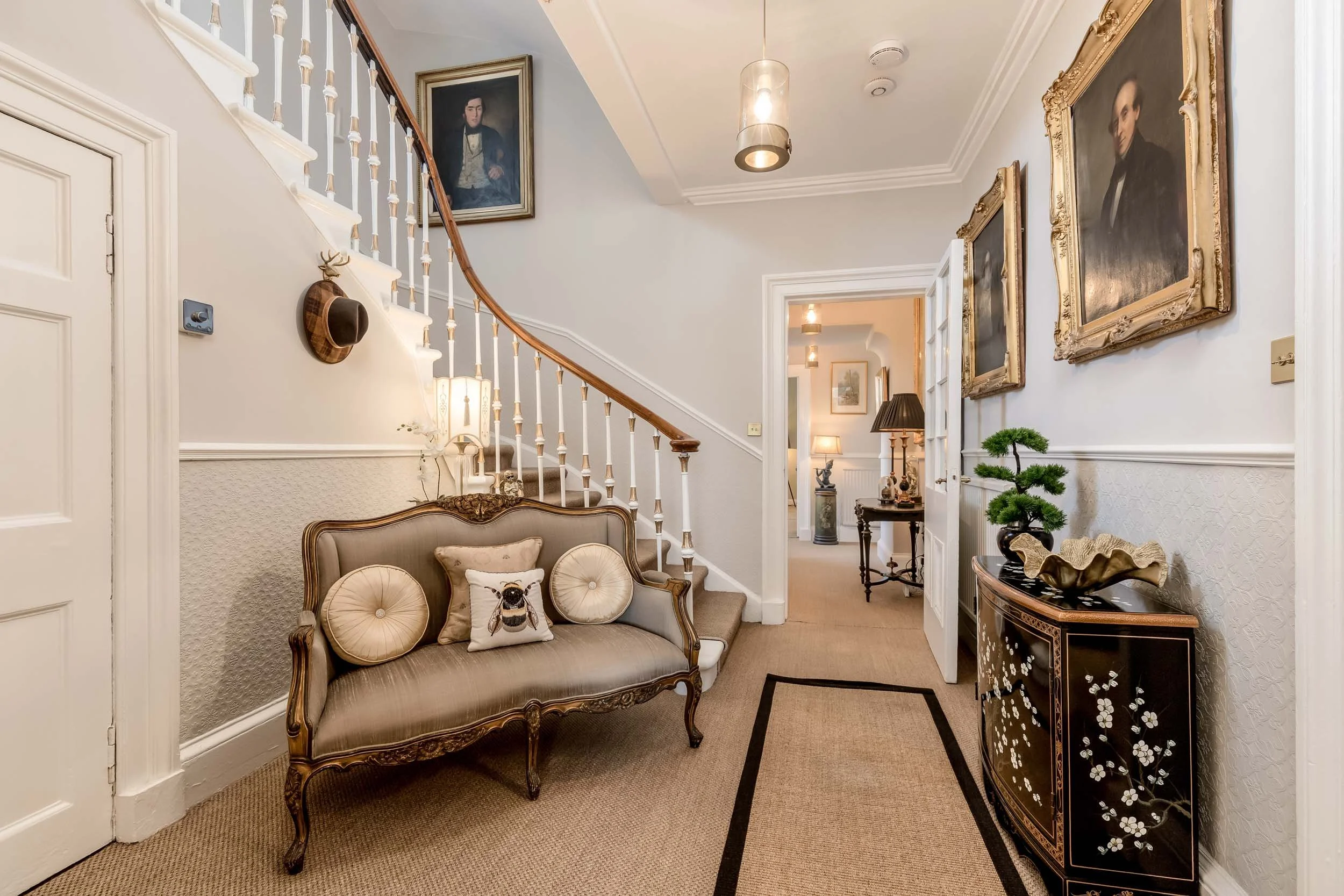 Interior of a hallway with decorative furniture and framed portraits on the walls, including a vintage-style sofa with pillows, a small black lacquered cabinet with floral design and a potted plant, and a glimpse into the next room with a table lamp 