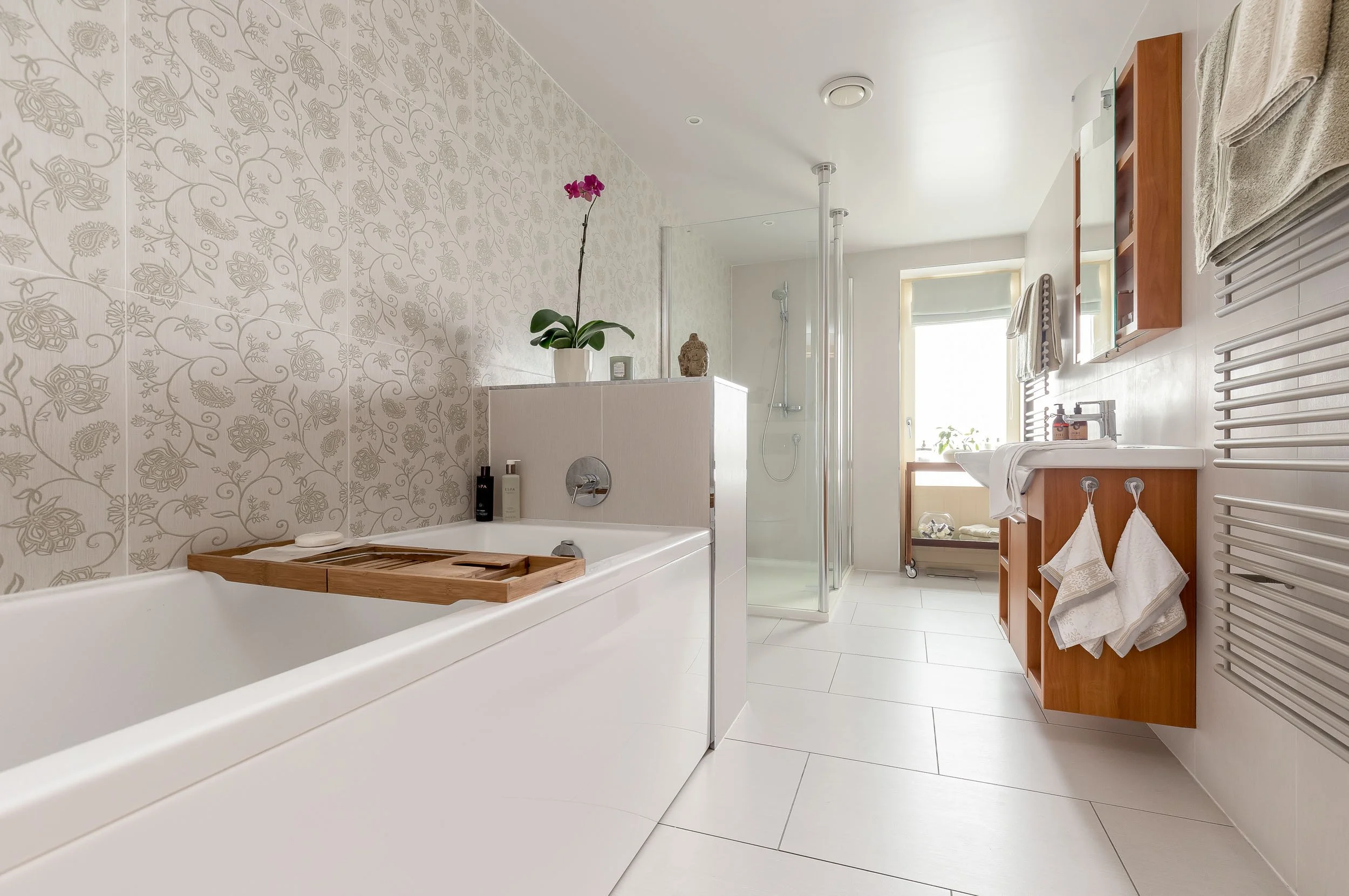 Bright modern bathroom with bathtub, shower, and wooden vanity with towels, plants, and toiletries.