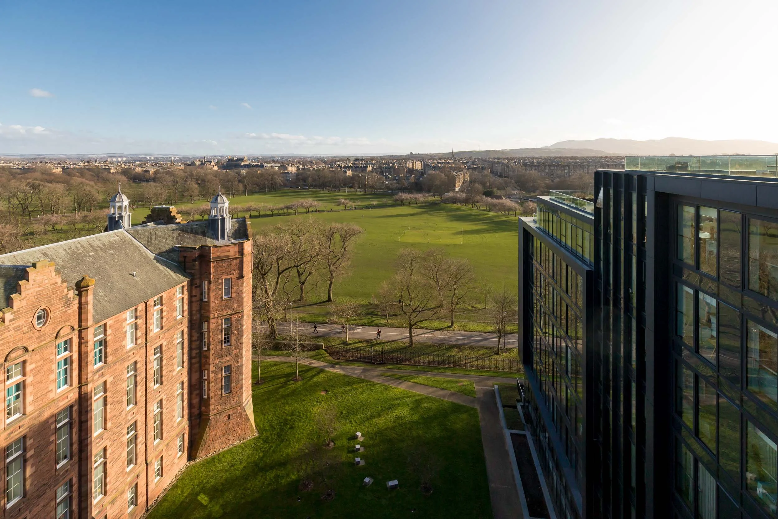 View of a park with green grass, trees, and walking paths, flanked by historic and modern buildings, with cityscape and mountains in the distance under a partly cloudy sky.