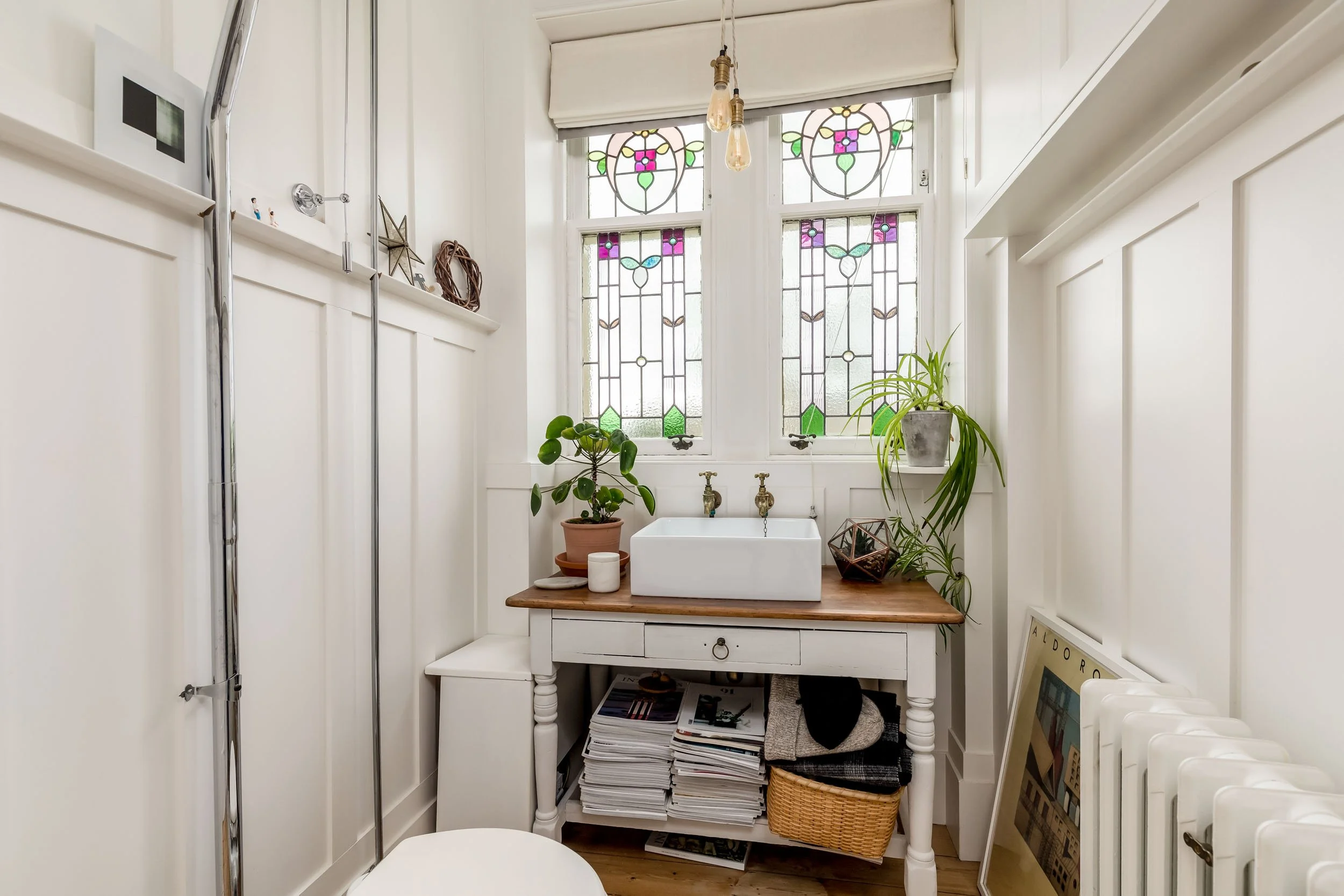 A small bathroom with white paneled walls, stained glass windows, a white ceramic vessel sink on a wooden top table, houseplants on either side, a pile of magazines and a basket underneath, and framed artwork leaning against the wall near a radiator.