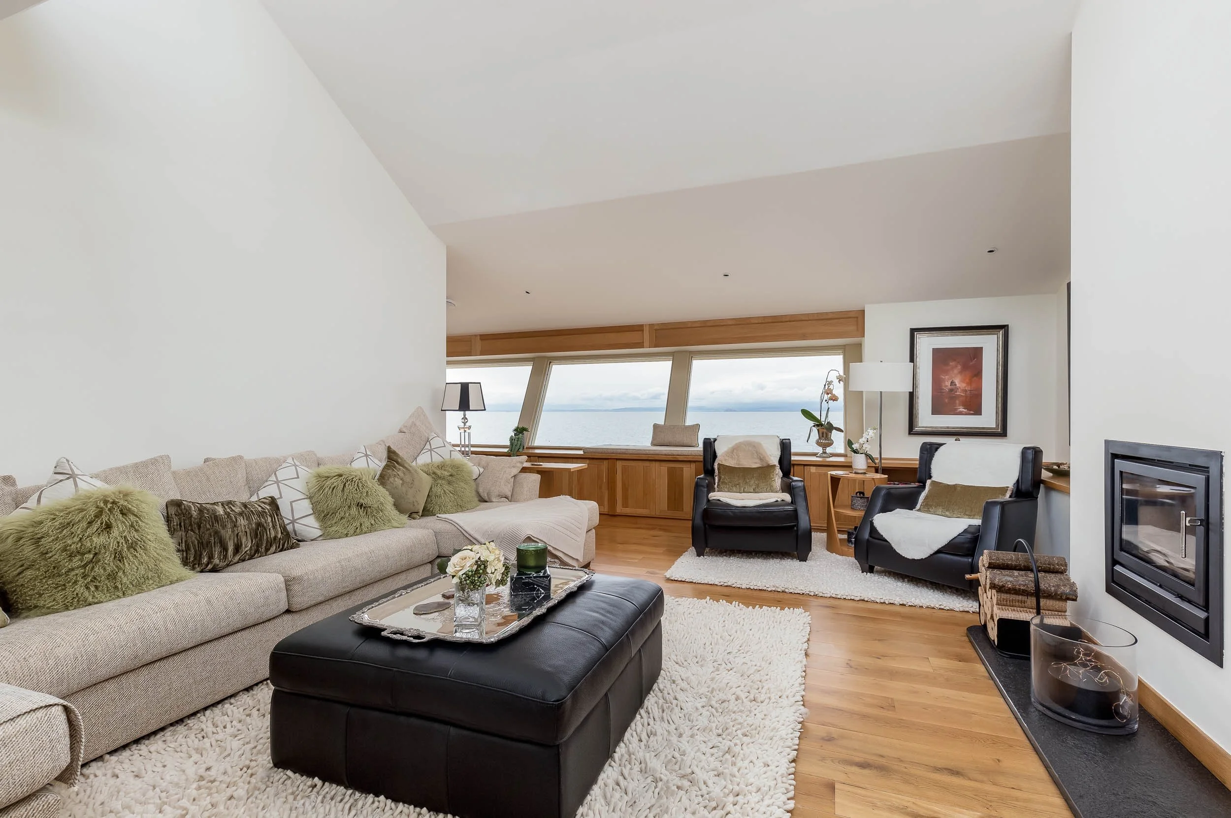 Modern living room with beige sofa, green and patterned pillows, black armchairs, a cream rug, wooden floors, decorative items, and large windows overlooking water.