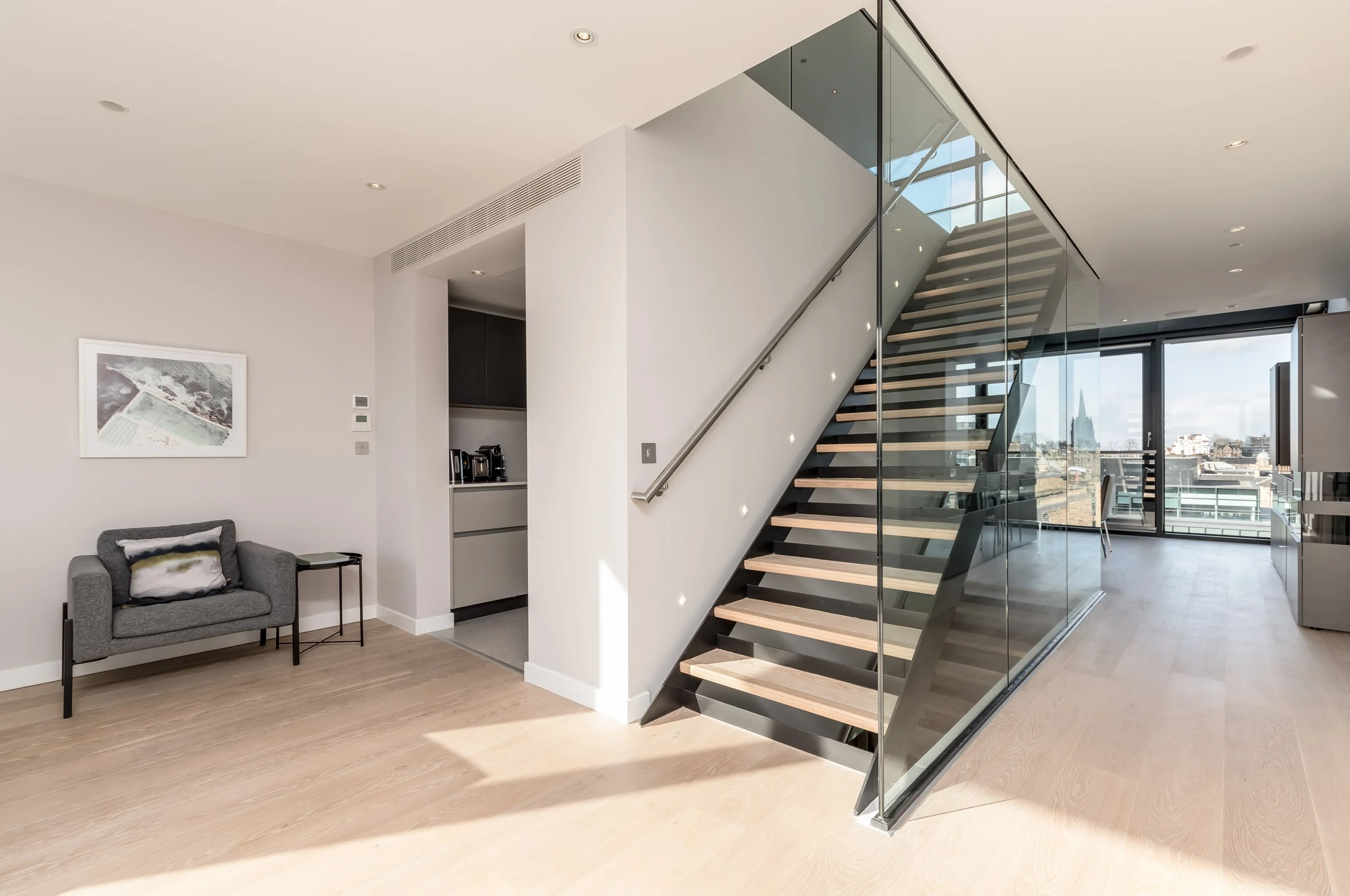Modern apartment living room with wooden stairs enclosed in glass, a gray sofa with a pillow, a small side table, a kitchen area with appliances, and large windows showing a city skyline.