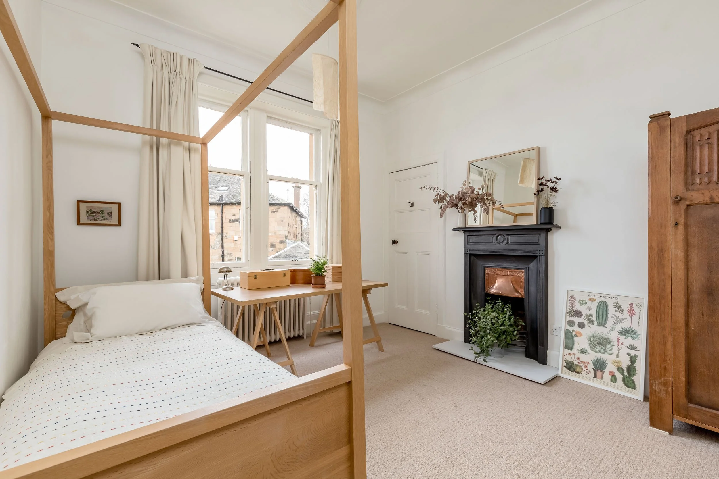 A cozy bedroom with a wooden bed frame, a desk by the window, a black fireplace with a plant on the hearth, a mirror above the fireplace, and vintage furniture.