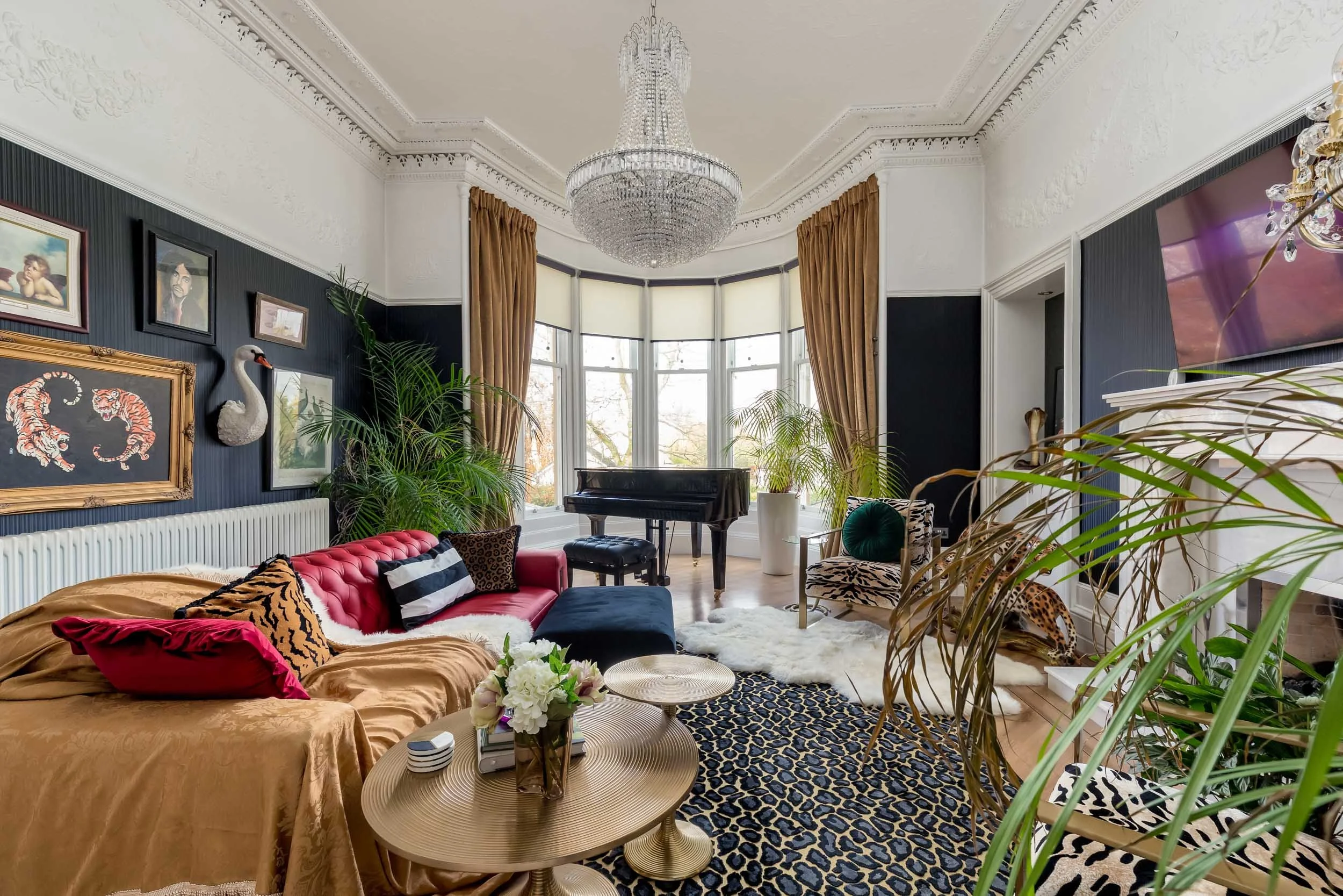 Luxury living room with dark walls, gold curtains, large bay window, velvet and animal print furniture, chandelier, and artwork.