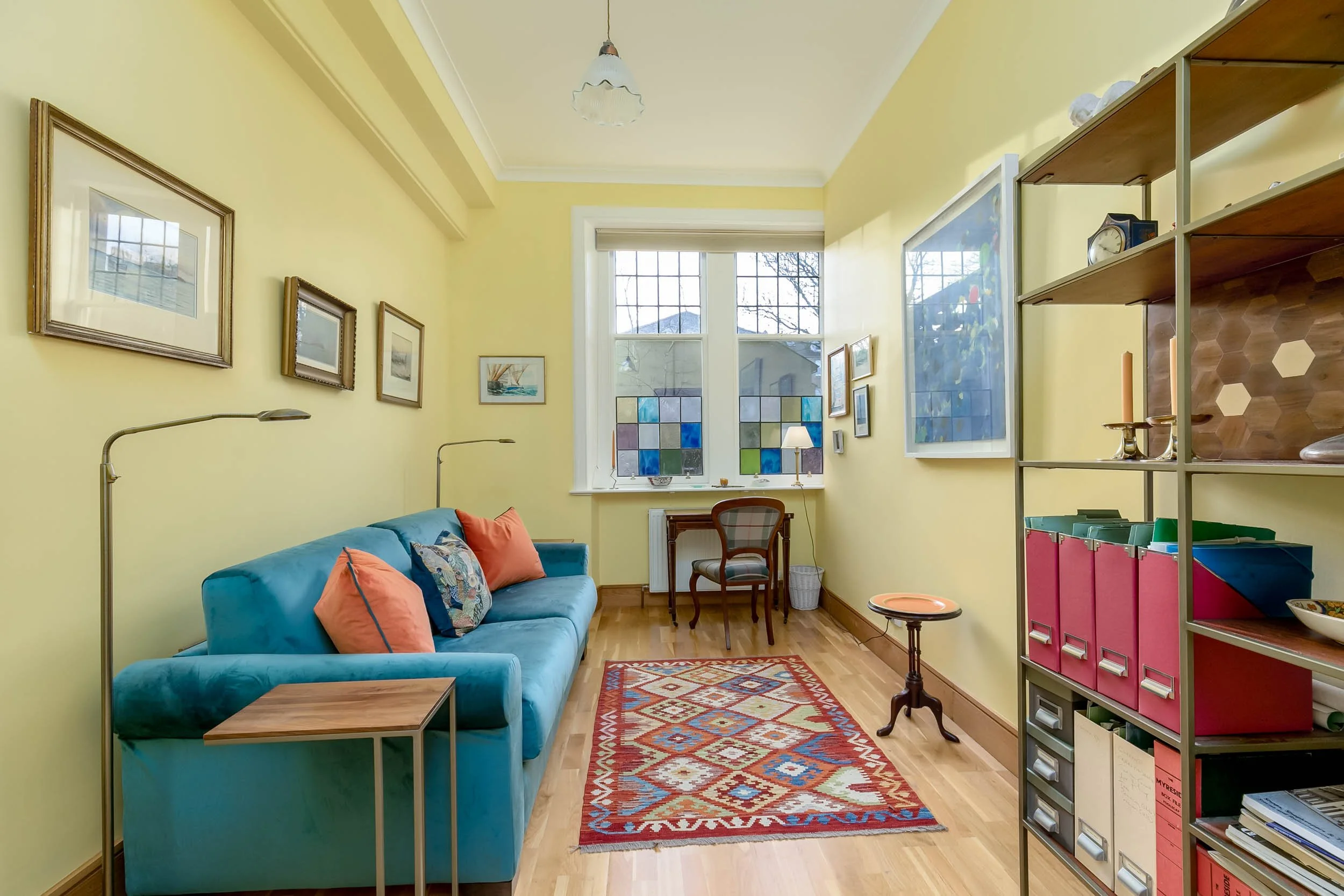 Bright yellow living room with wooden floors, blue sofa with orange pillows, colorful patterned rug, bookshelf, and window with stained glass panels