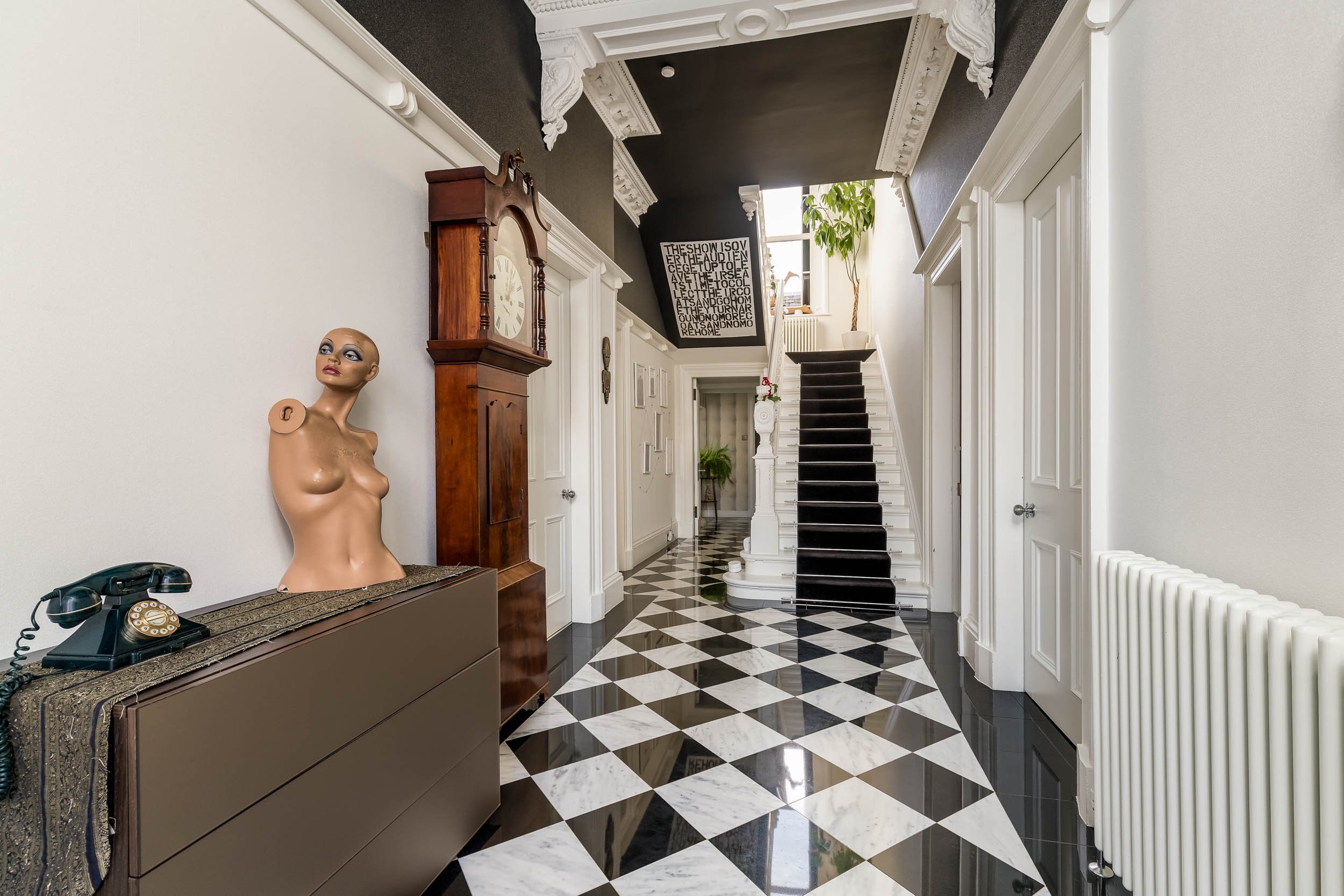 Elegant foyer with black and white checkered marble floor, vintage mirror on wall, wooden grandfather clock, mannequin bust, black staircase with white balusters, potted plants, and decorative molding.
