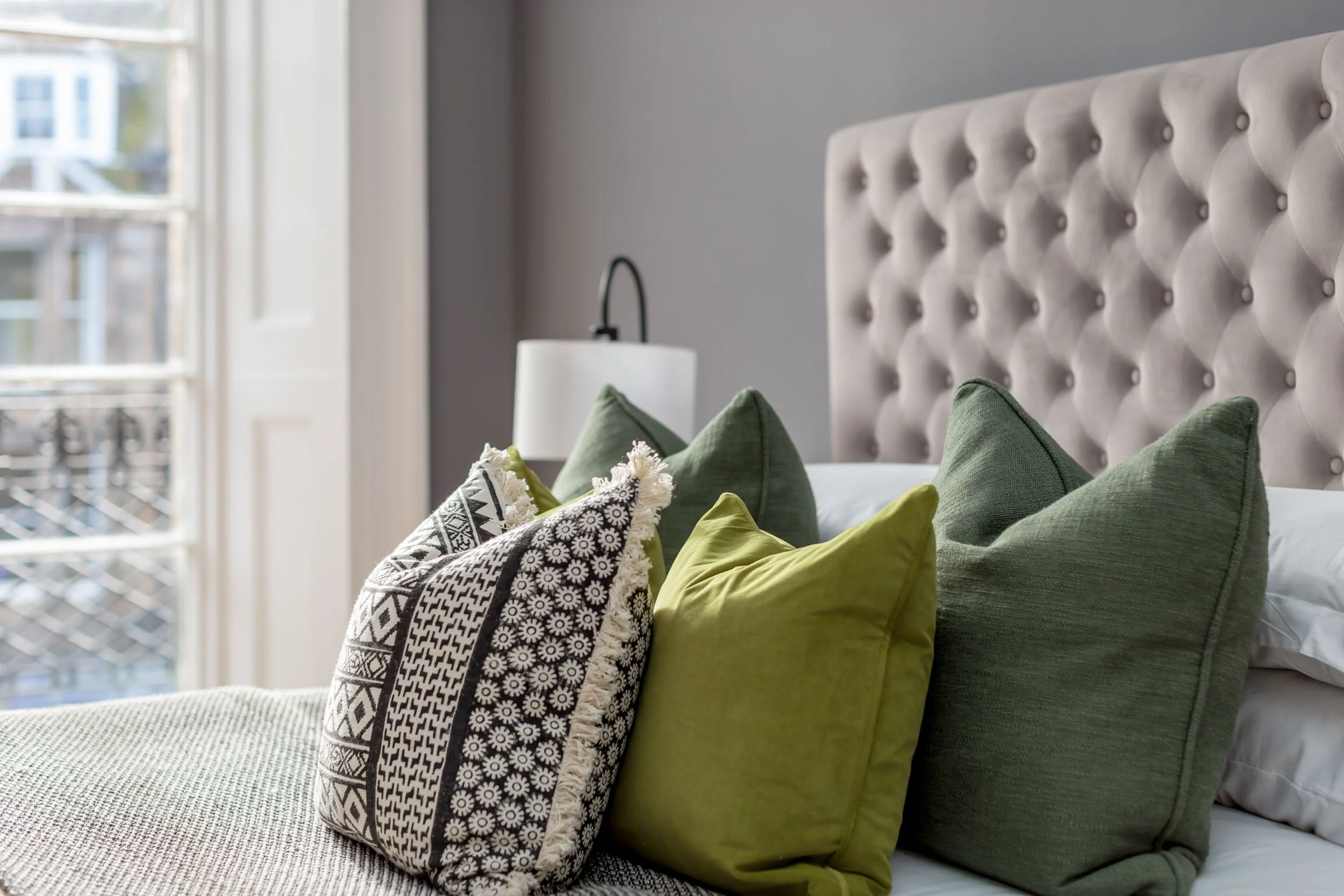 A neatly made bed with multiple green and patterned pillows arranged against a padded beige headboard, next to a window with white curtains and a lampshade
