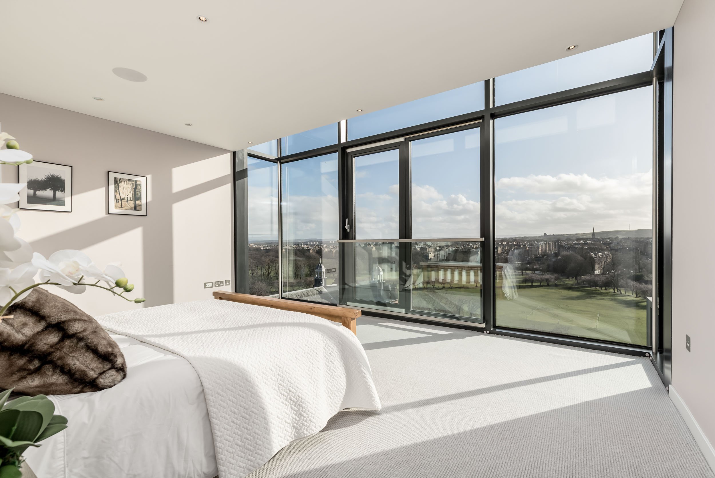 Modern bedroom with large floor-to-ceiling windows showing cityscape and green field outside, white bed with a brown fur pillow, and minimal wall art.