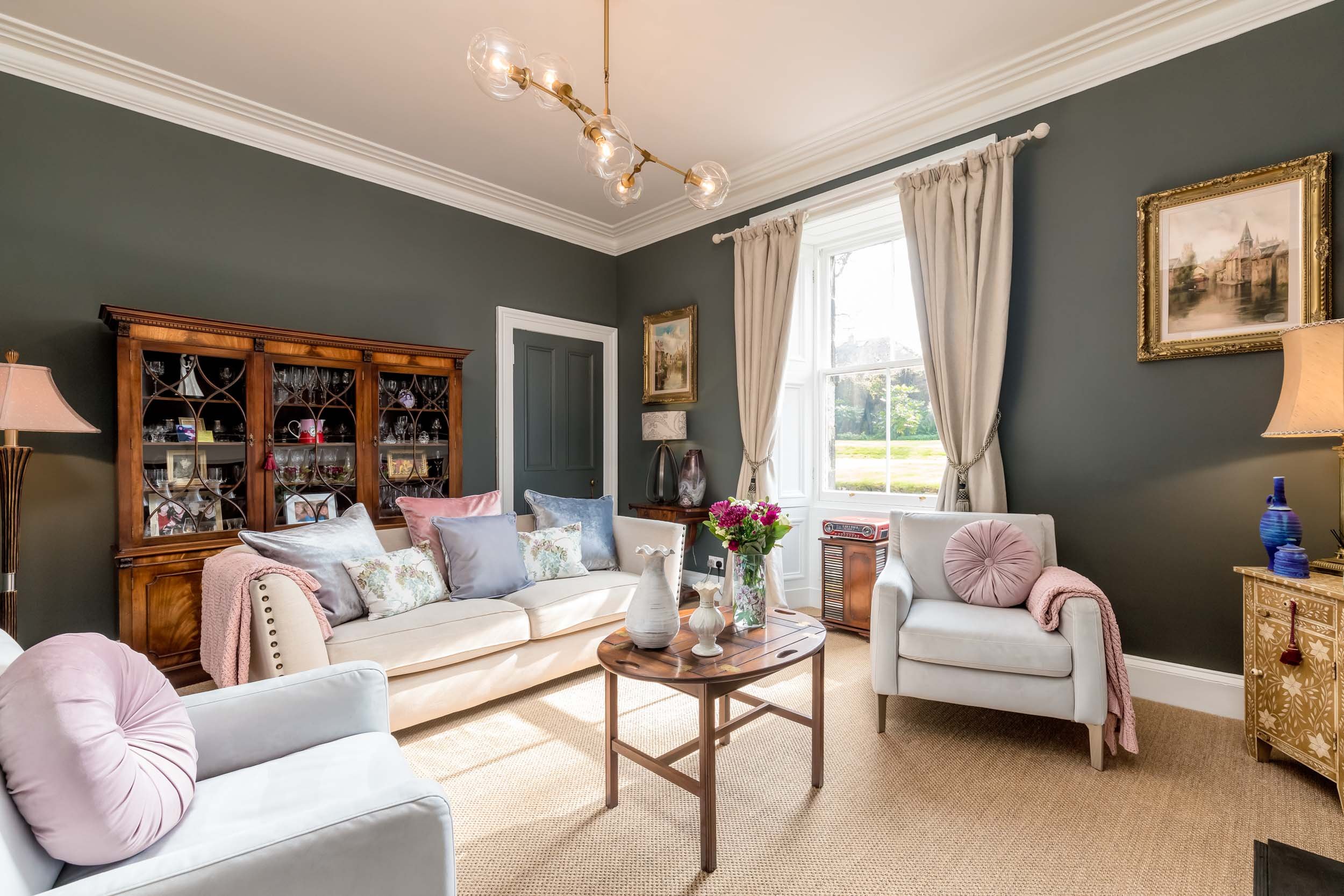 Living room with dark green walls, cream sofa and armchair with pink pillows, a wooden china cabinet, a round wooden coffee table, floral arrangements, framed paintings, and beige curtains, lit by a modern chandelier.