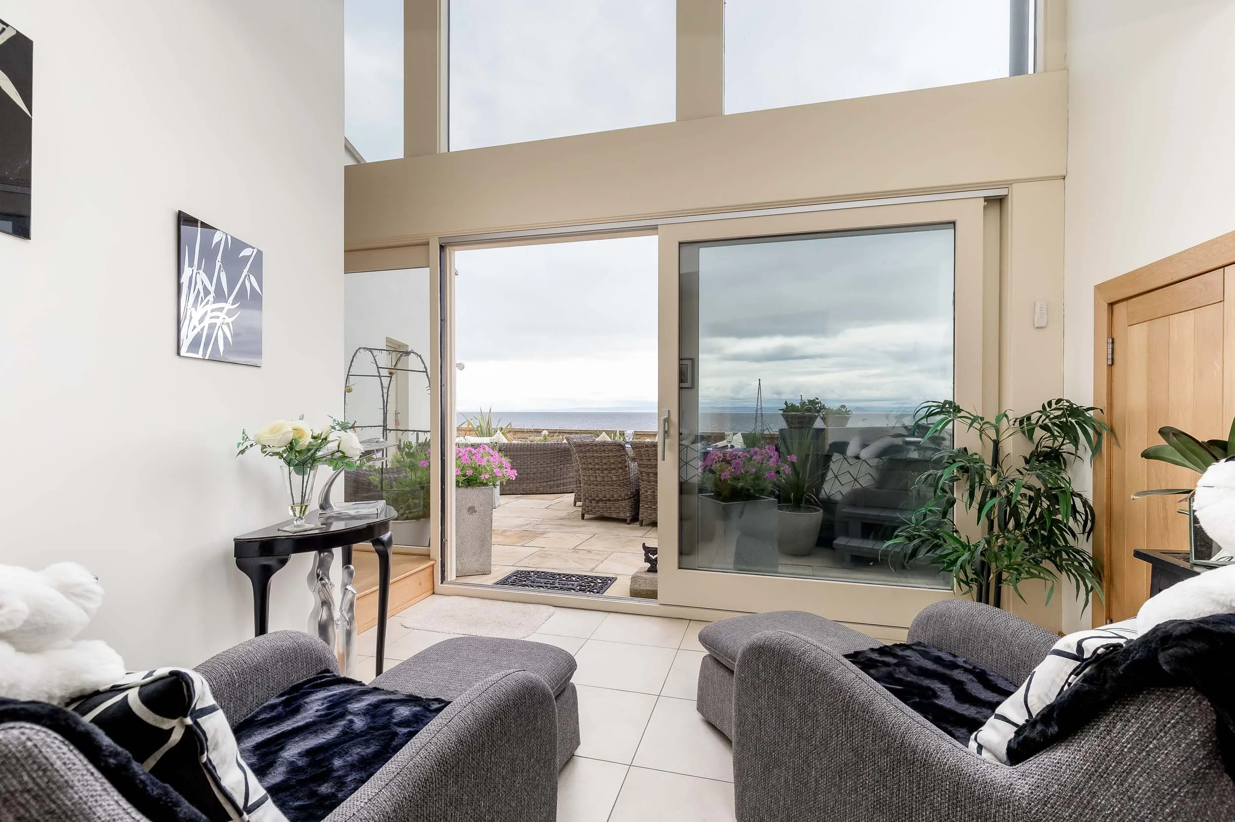 Living room with two gray armchairs, a small console table with a flower vase, sliding glass door leading to a balcony with outdoor furniture and potted plants under cloudy sky.