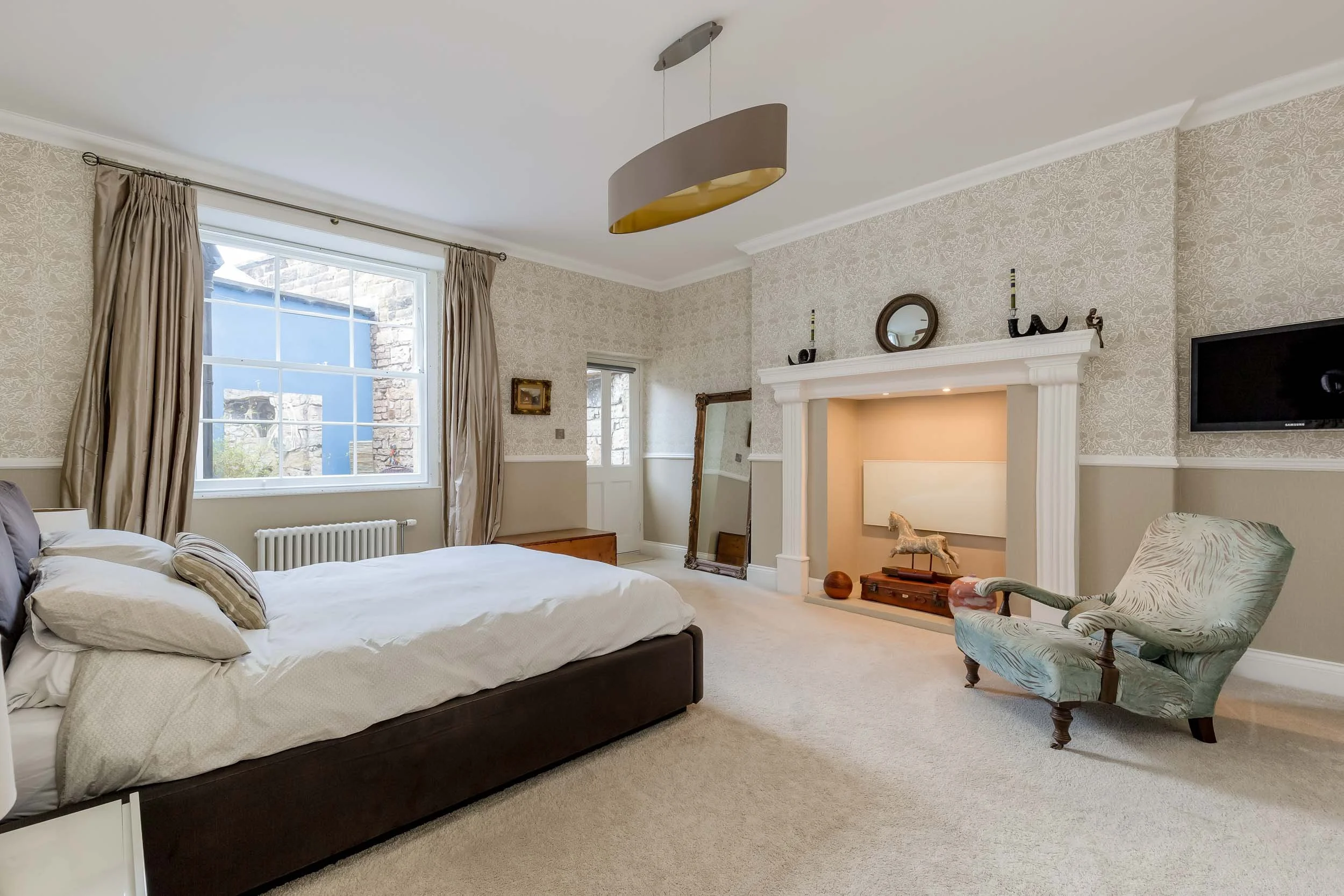 A bedroom with a large bed, window with curtains, a decorative fireplace mantel with artifacts, a vintage mirror leaning against the wall, an armchair, and a wall-mounted TV.