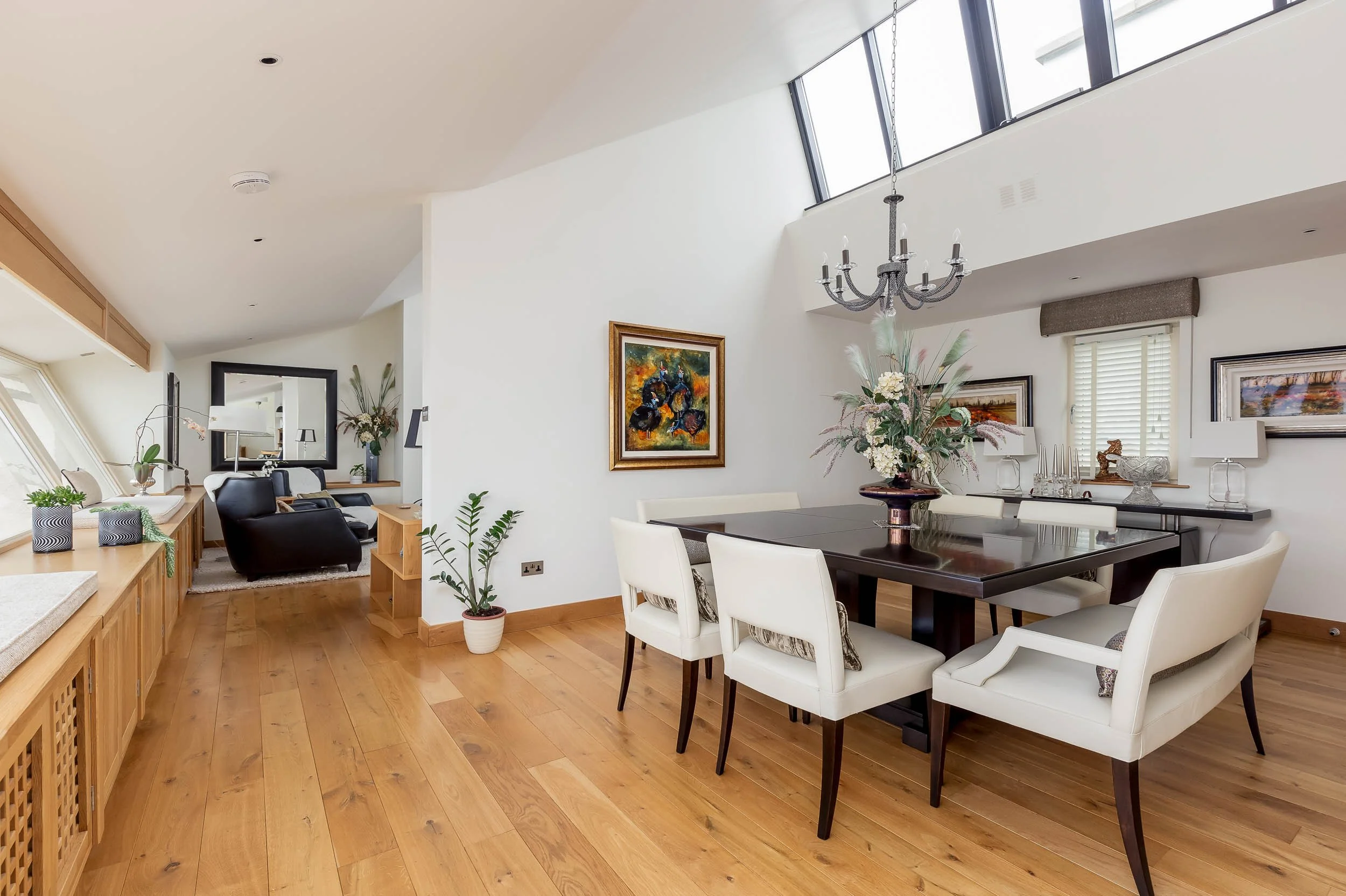 Dining area with a dark wood table and white chairs, decorated with a floral centerpiece. Adjacent to a living room with two black armchairs and a mirror, with artwork on the walls and windows letting in natural light.
