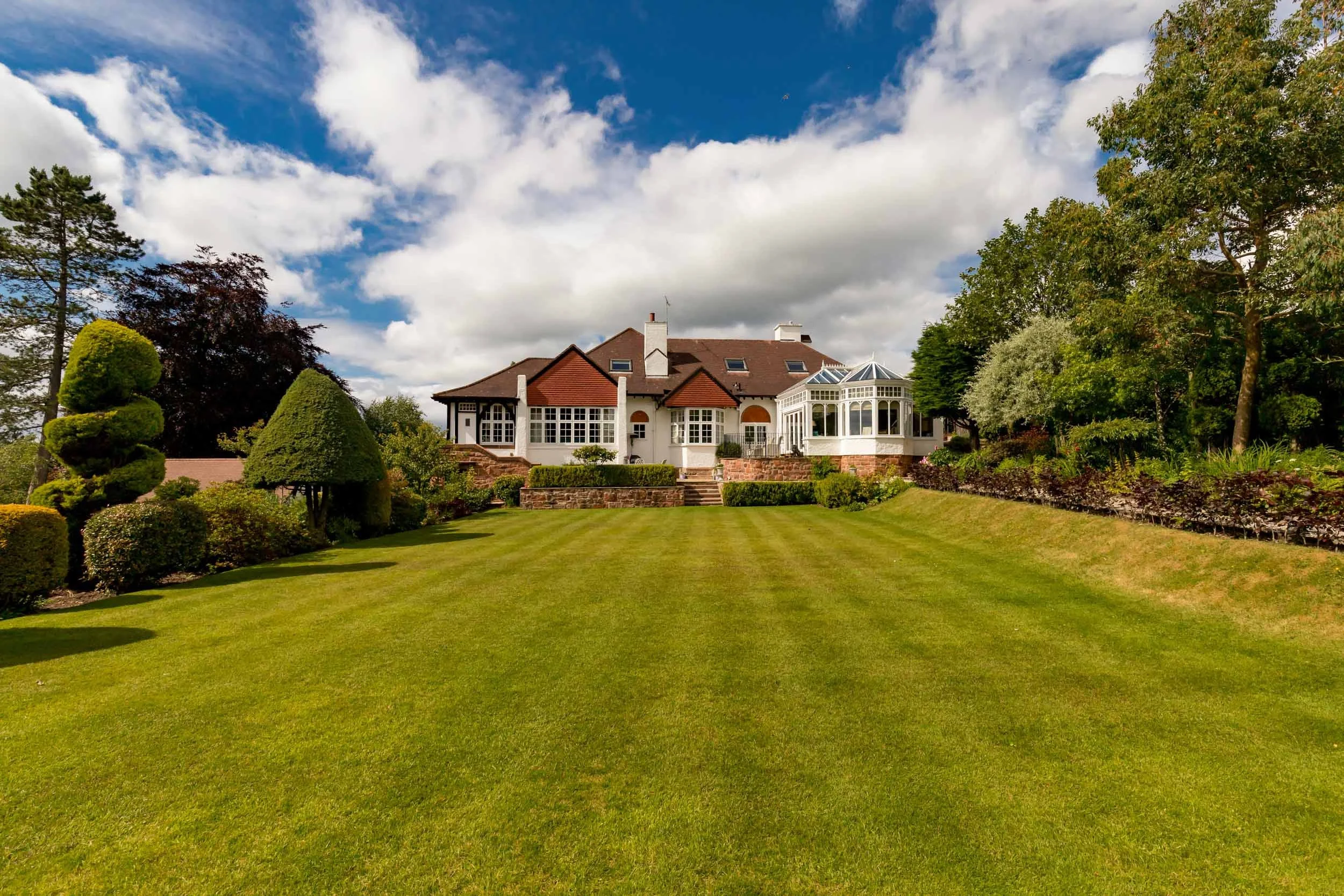 Large house with a brown roof and white walls, surrounded by a well-maintained lawn and lush green trees and bushes.
