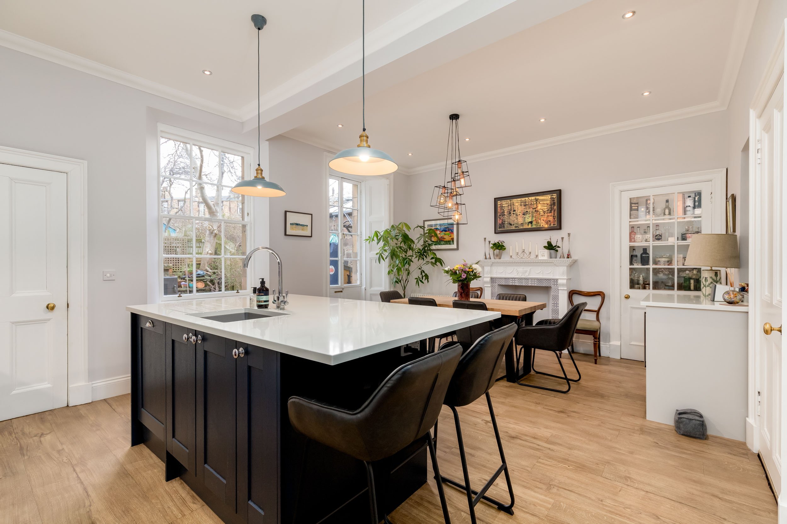 Modern kitchen and dining area with white walls, large windows, a black kitchen island with white countertop, pendant lights, and a white fireplace with artwork and decorative items.