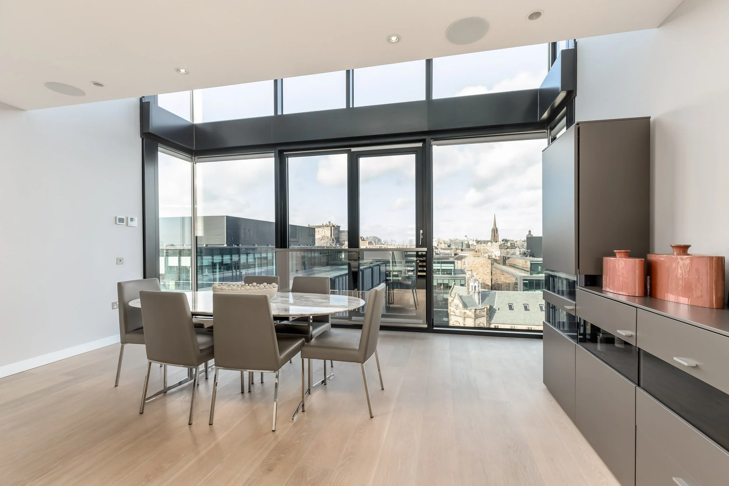 Modern dining room with large floor-to-ceiling windows, a round dining table with six beige chairs, and a gray cabinet with pink decorative vases on top.