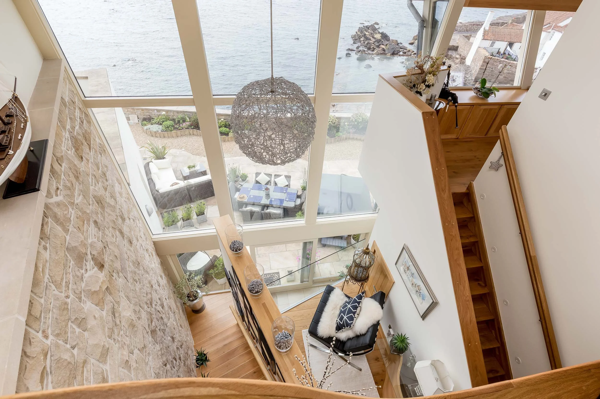 A picture of the interior of a multi-story house, taken from the upper level. The house has large glass windows overlooking a body of water and a patio with outdoor furniture. Inside, there are wooden floors, a black armchair with a white fluffy pill