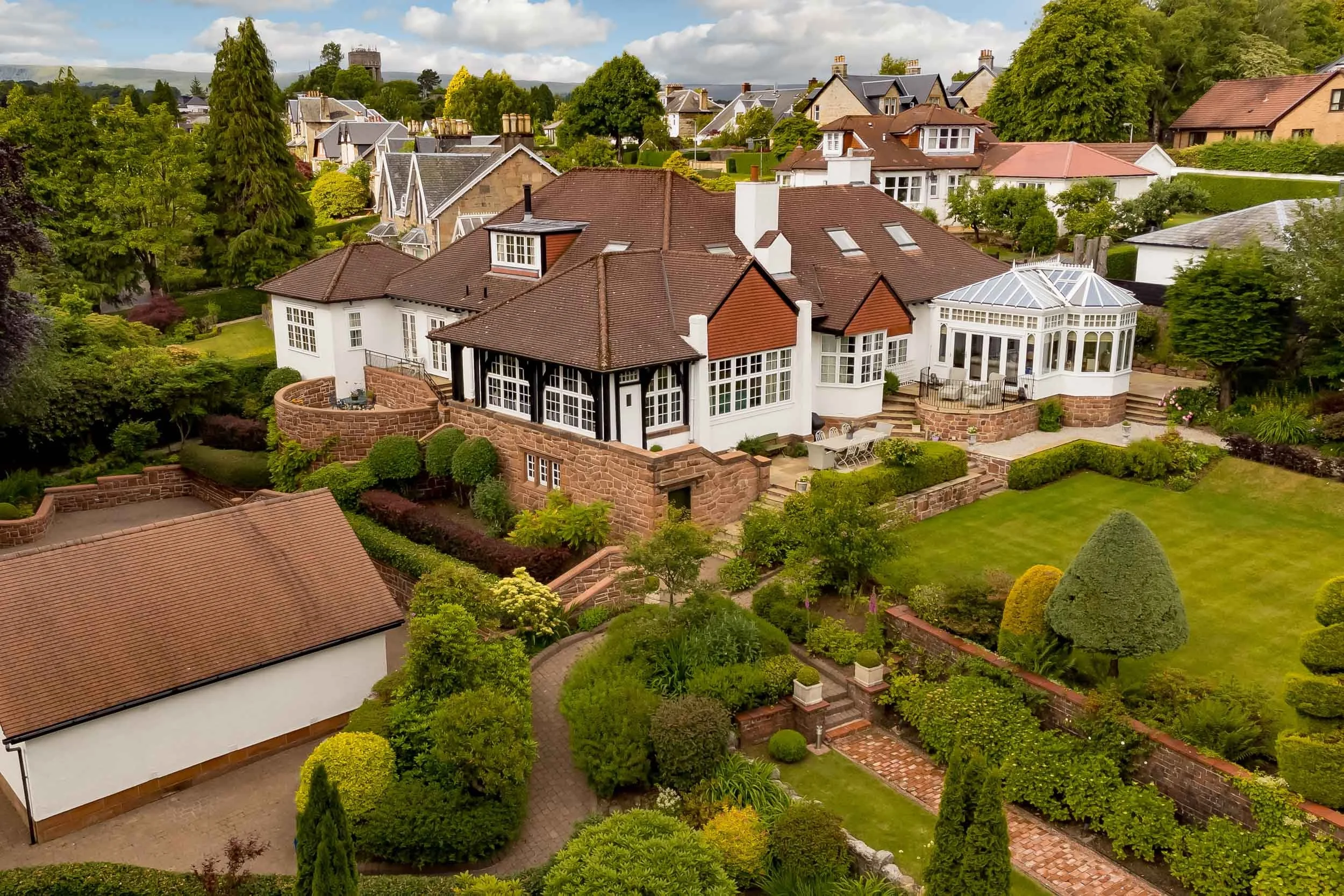 A large, multi-story house with a red and white exterior, surrounded by a lush, well-manicured garden with various trees and shrubs. The house features multiple roofs, a glass conservatory, and outdoor seating areas.