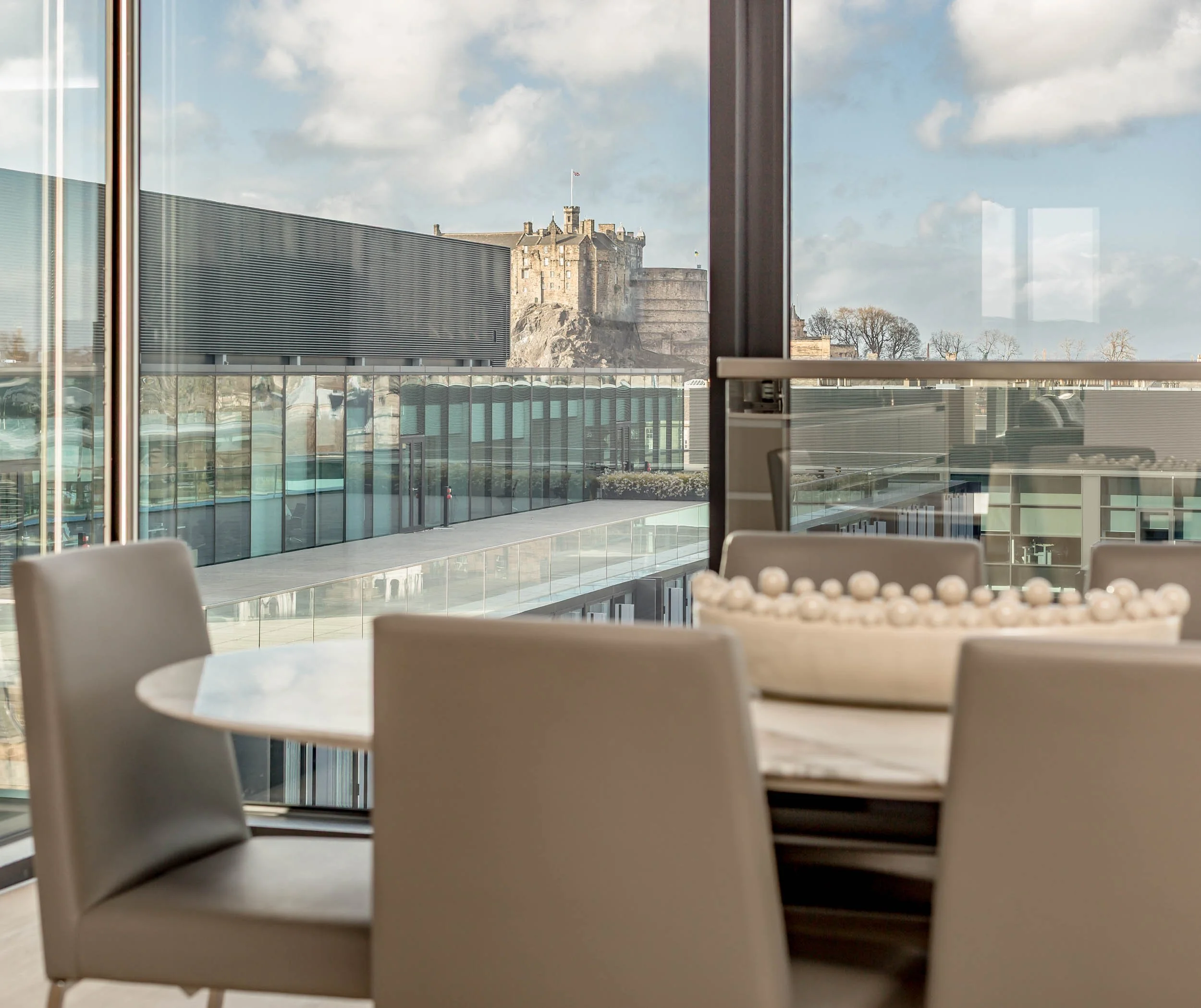 Interior view of a modern building with a round table and beige chairs, with a large window showing a historic castle on a hill outside.