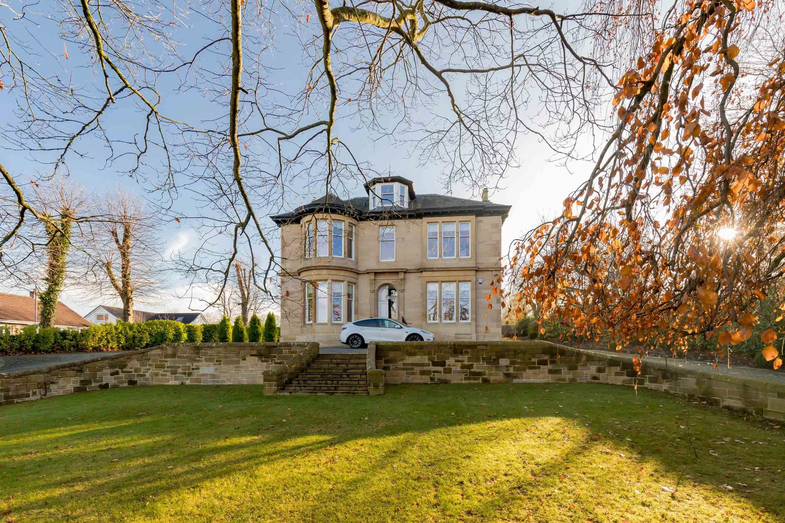 A large, two-story historic house with a bay window and dark roof, situated behind a stone wall with stairs, a white car parked at the entrance, leafless trees in the background, and some autumn-colored tree branches on the right, with the sun shinin