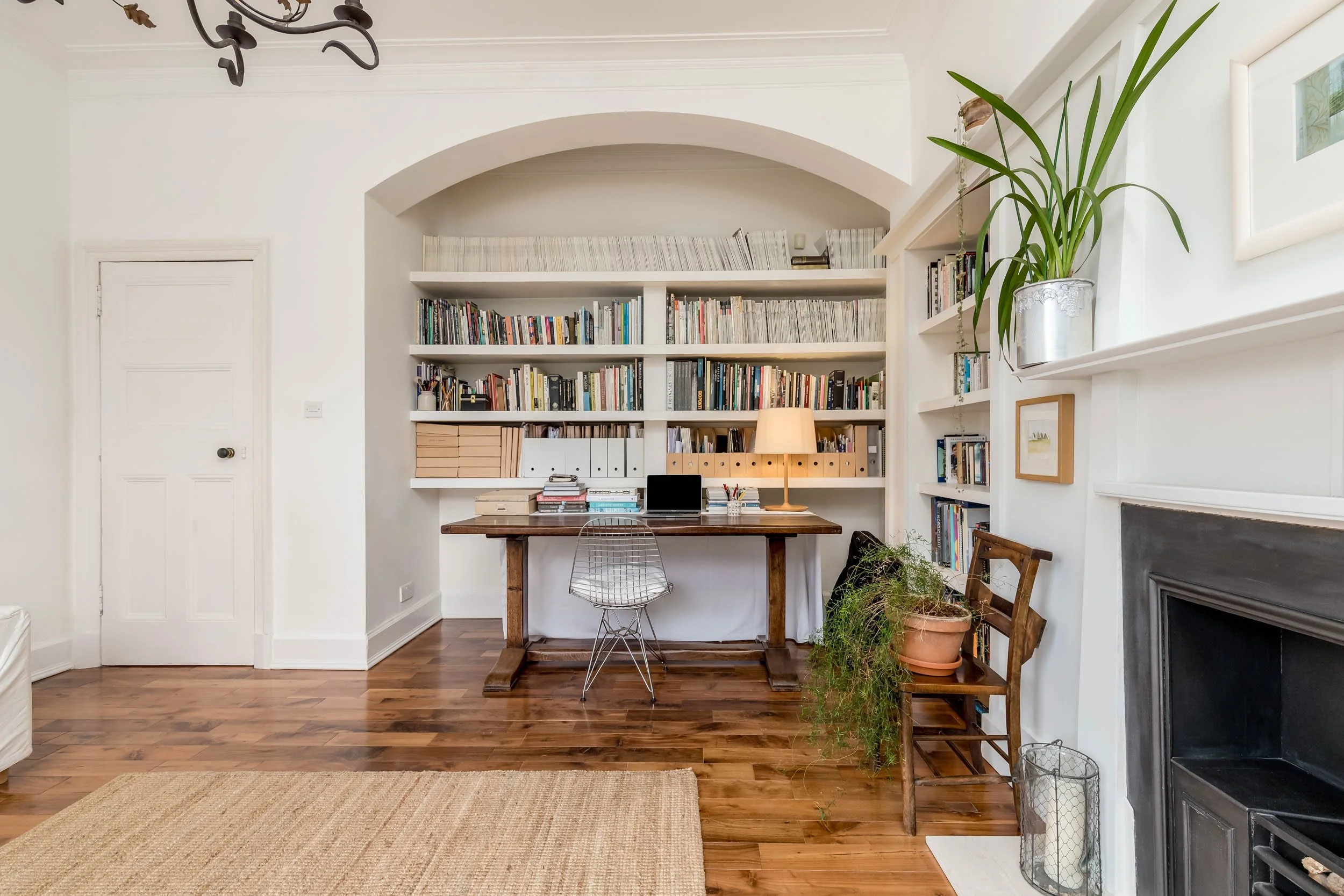 A cozy home office with a wooden desk, a wire chair, and a bookshelf filled with books and binders. There are potted plants on the right side near the fireplace, and a lamp on the desk provides warm lighting. The room has white walls, a wooden floor,