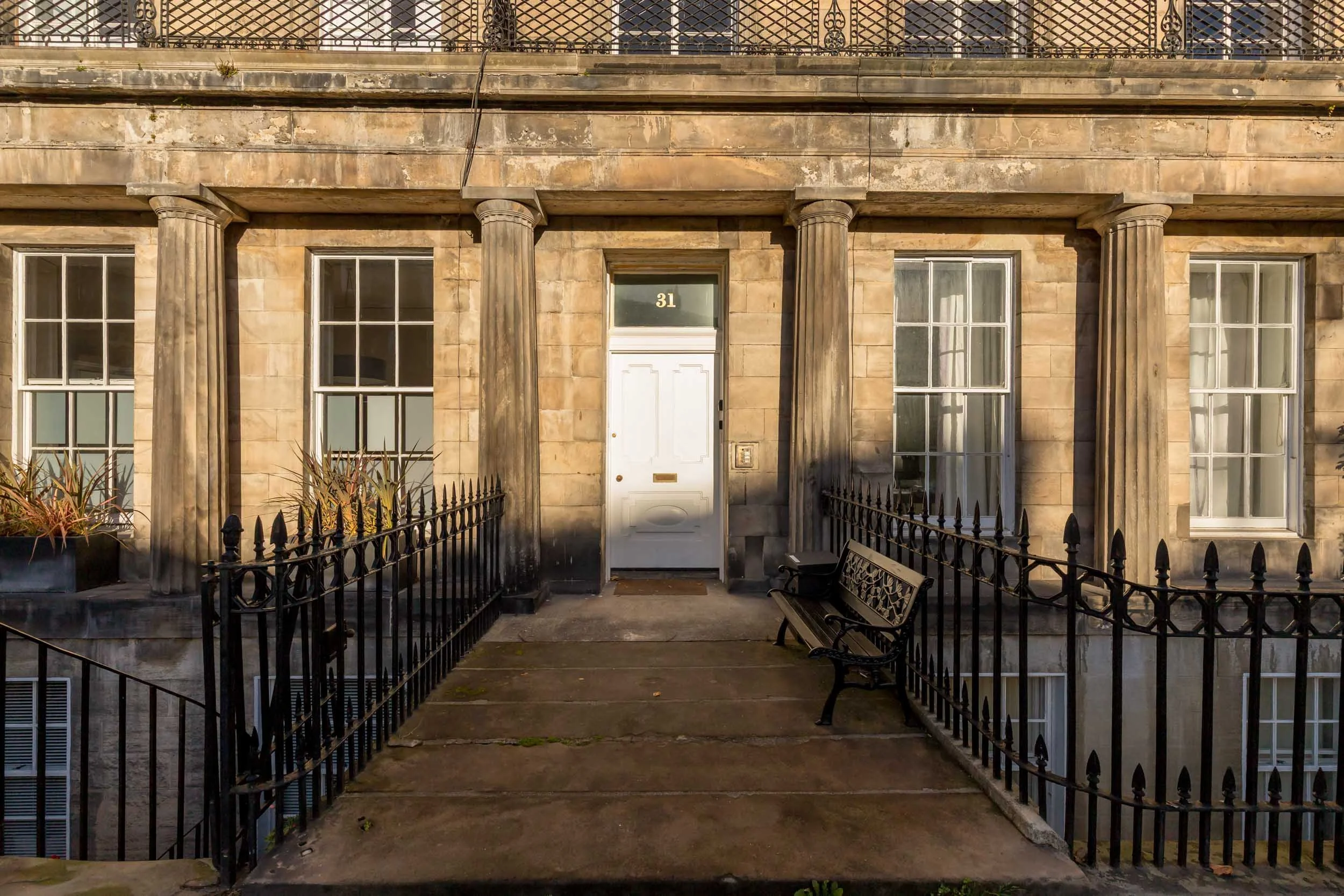 Front entrance of a classic apartment building with six columns, three on each side of the central white door labeled 31, black iron railing, a metal bench, and two large windows with white frames.