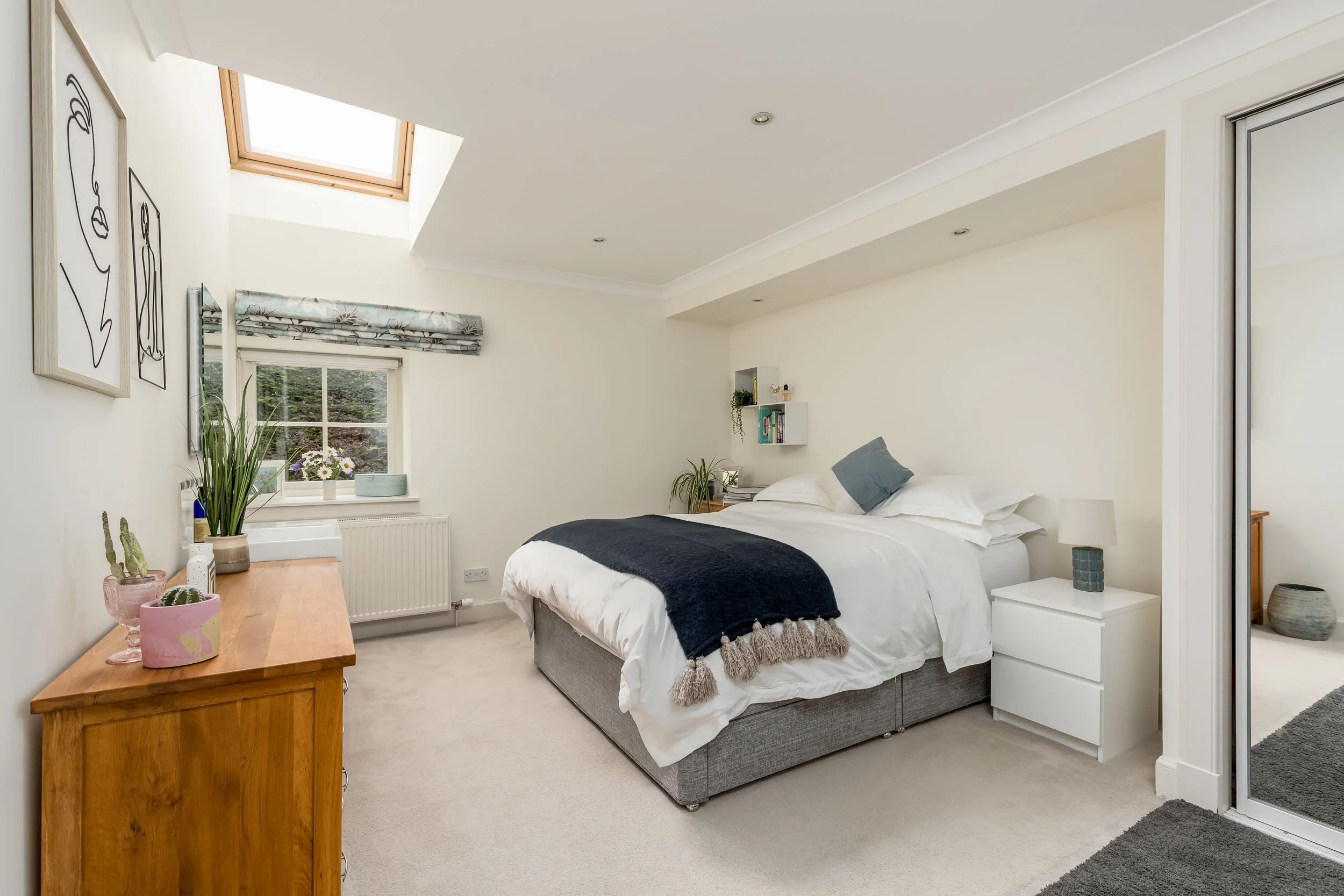 A bedroom with a skylight, a window with a floral curtain, a double bed with white bedding, a black blanket, and a blue accent pillow. There are wall art pieces, potted plants, a wooden dresser with plants, and a small bedside table with a lamp. A mi