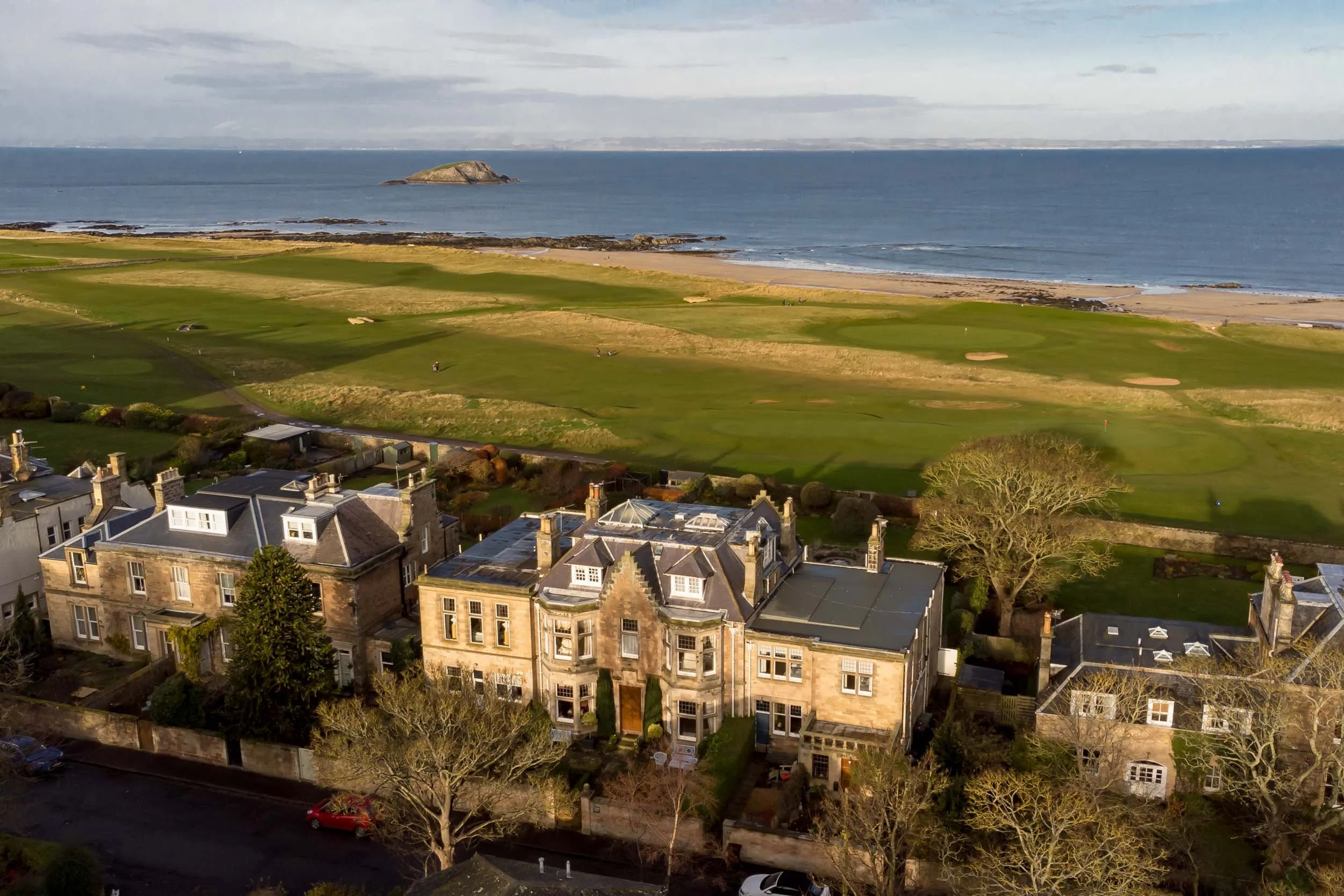 Aerial view of a residential area with large houses, a green golf course, and a beach with rocks and the ocean in the background.
