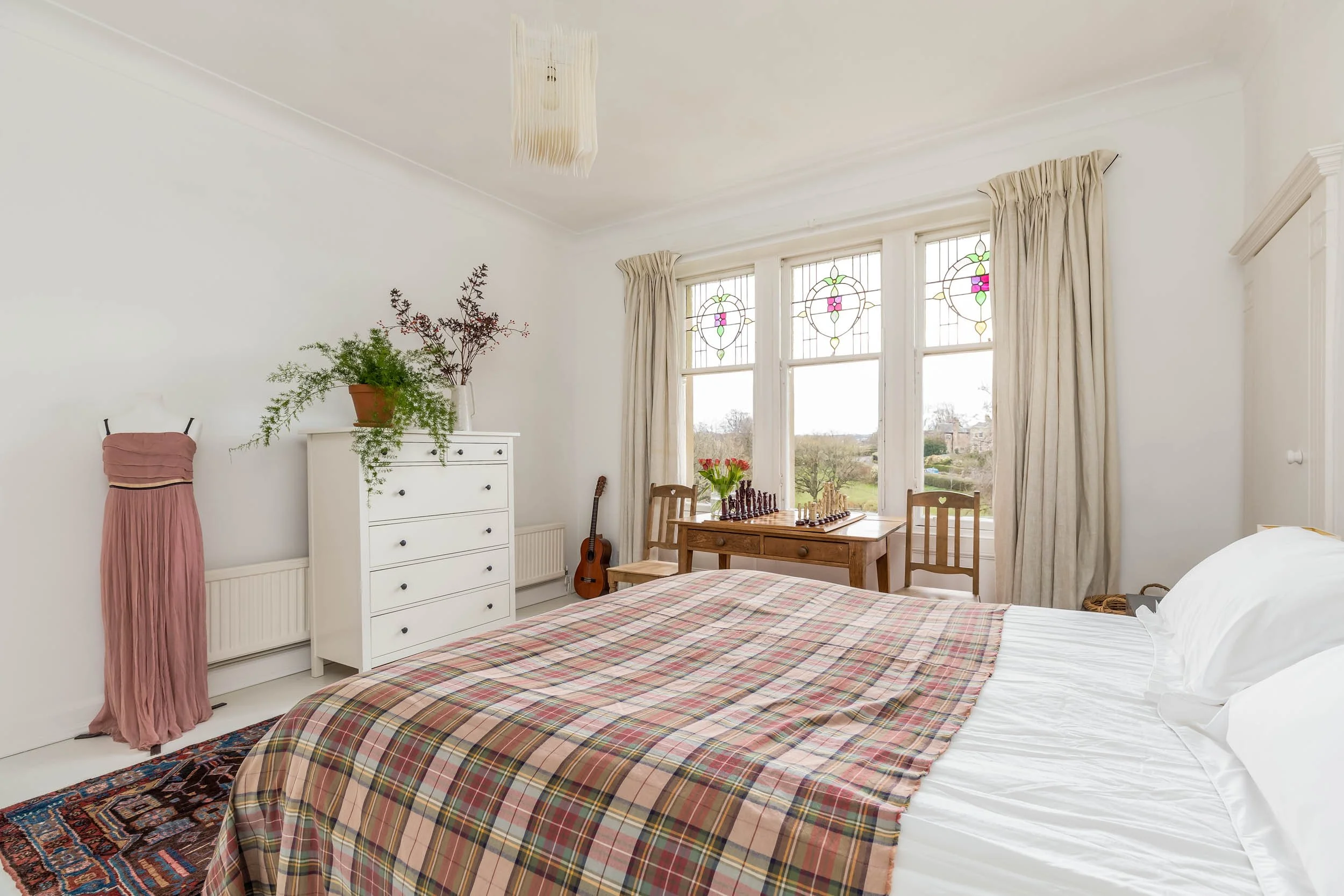 Bright bedroom with white walls and large stained glass windows, featuring a bed with a plaid quilt, a wooden table with flowers and chess, a white dresser with plants, and a dress hanging on the wall.